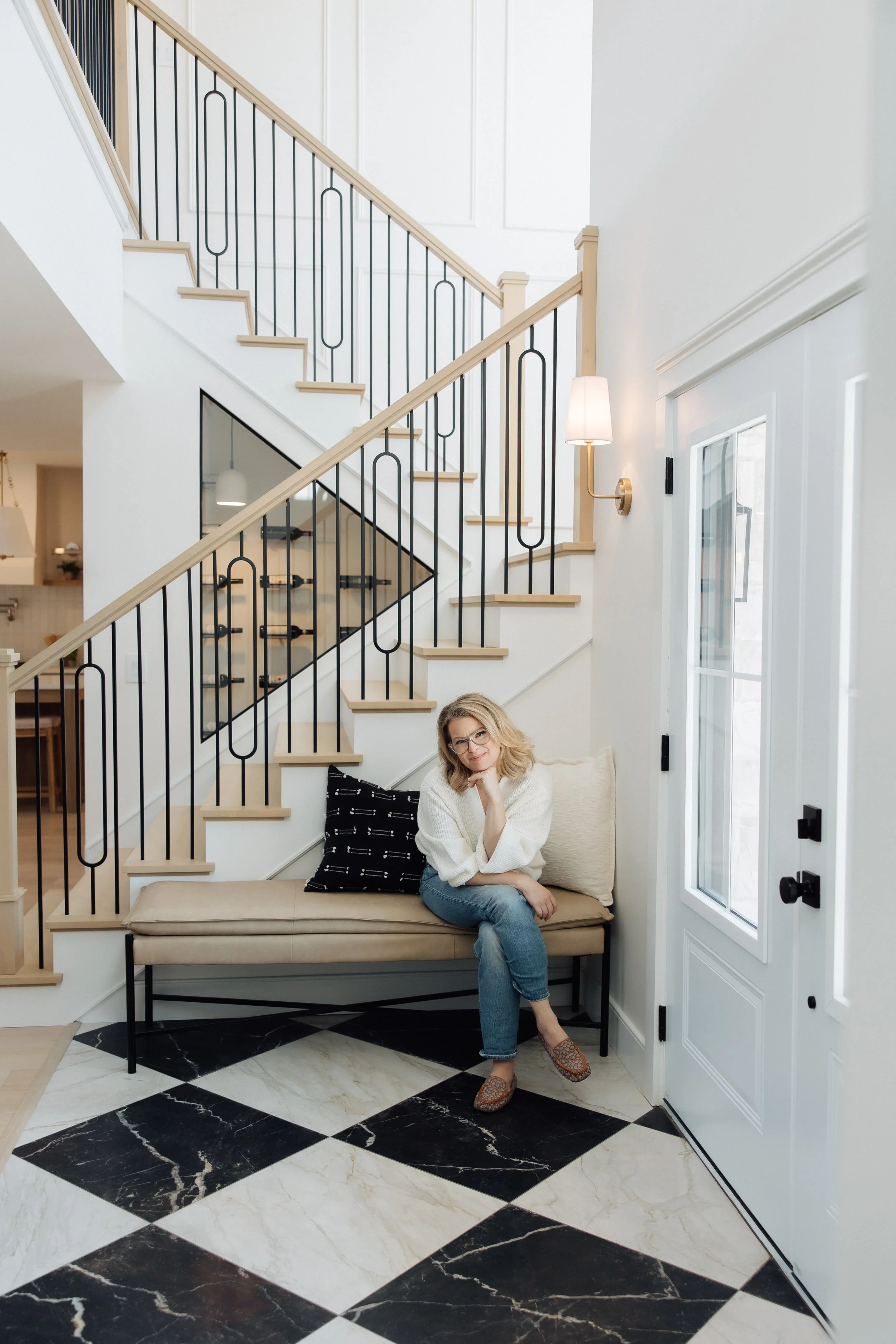 A woman with blonde hair, glasses, and a white sweater, sitting on a beige bench with black and white pillows in front of a staircase with black metal railing and light wood handrails, beside a glass door and a white wall lamp.