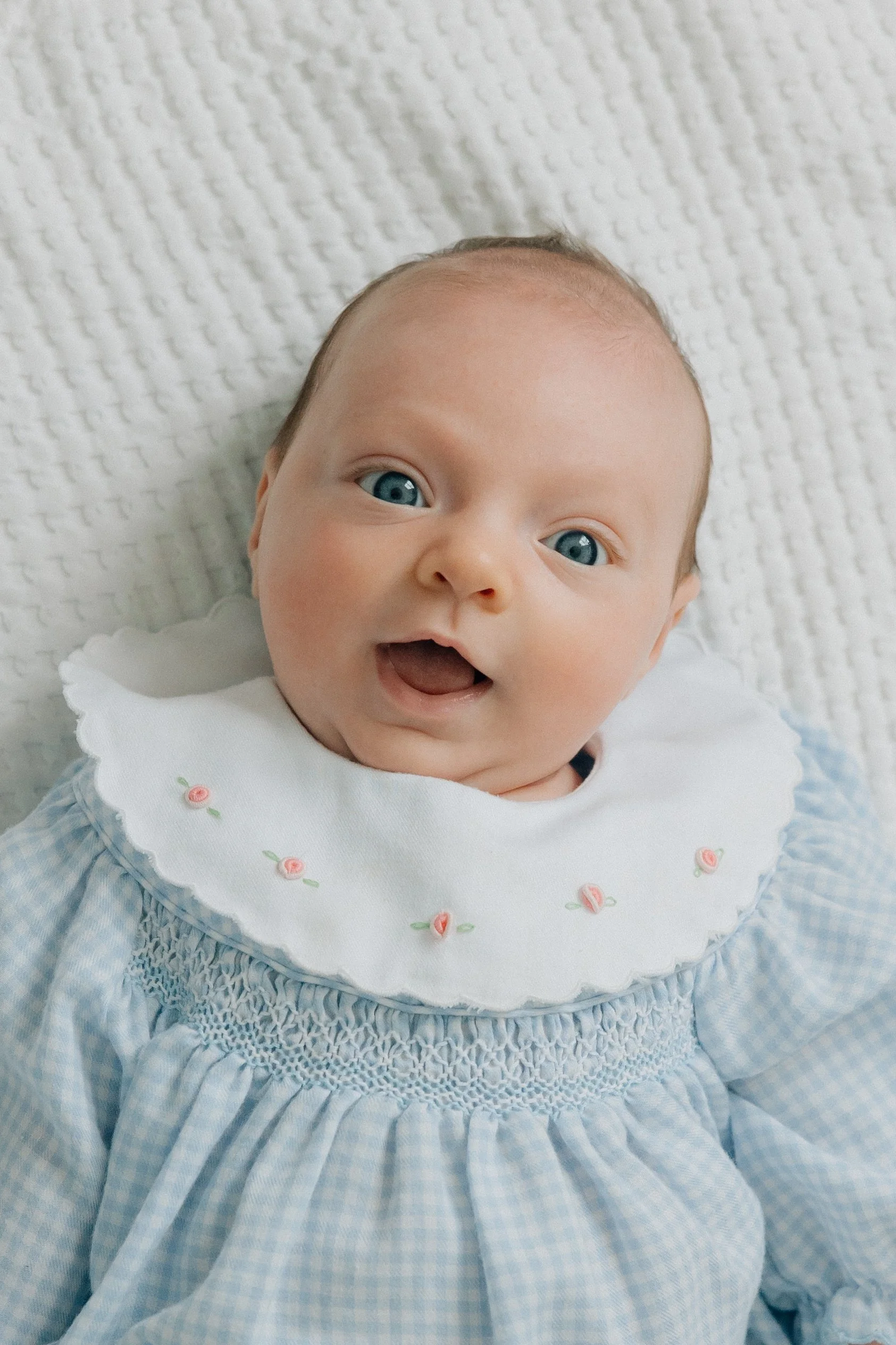 Close-up of a smiling baby with blue eyes, wearing a white bib with pink embroidery, a blue gingham dress, and a blue smocked dress, lying on a textured white blanket.