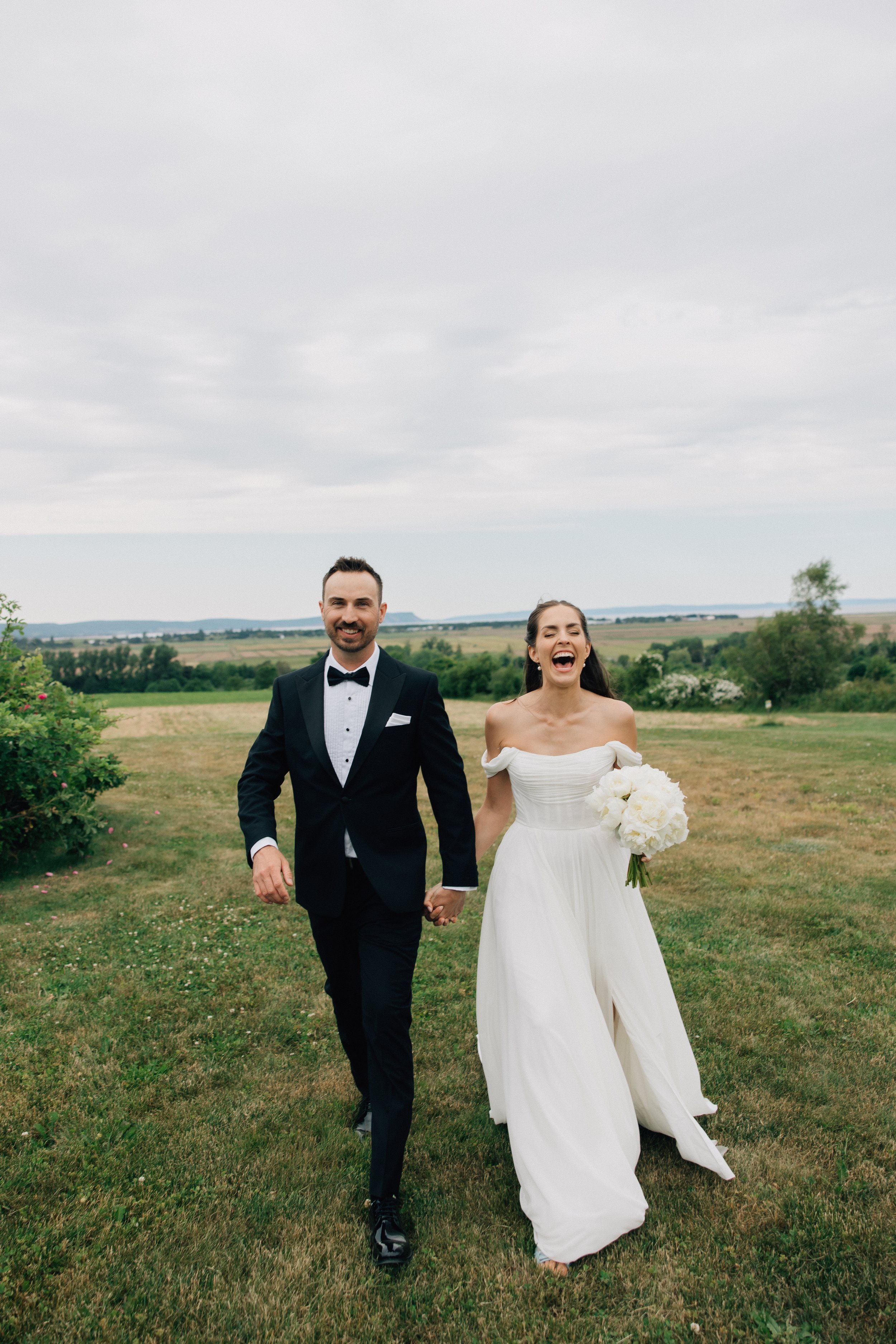 A smiling bride and groom holding hands and walking outdoors on a grassy field on a cloudy day. The groom is wearing a black tuxedo with a bow tie, and the bride is in a white strapless wedding dress, holding a bouquet of white flowers.