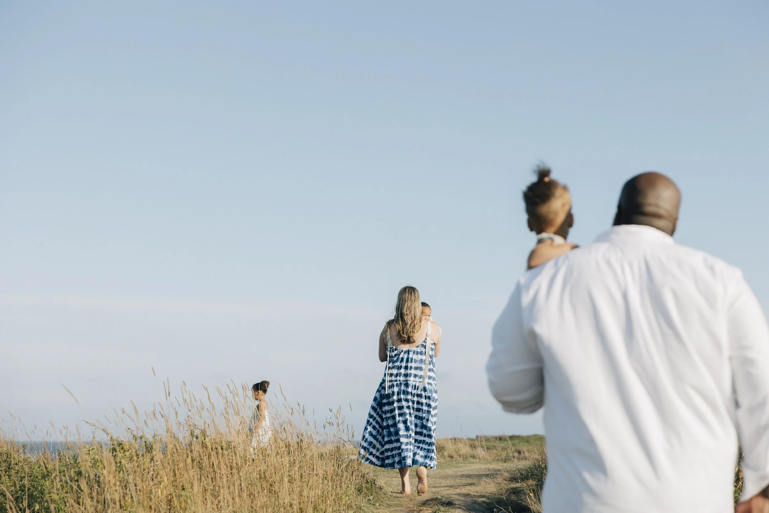 A group of people, including a man with a shaved head and a young woman with long hair, walking through a grassy field on a clear day. In the background, there are additional people, and the sky is blue with no visible clouds.