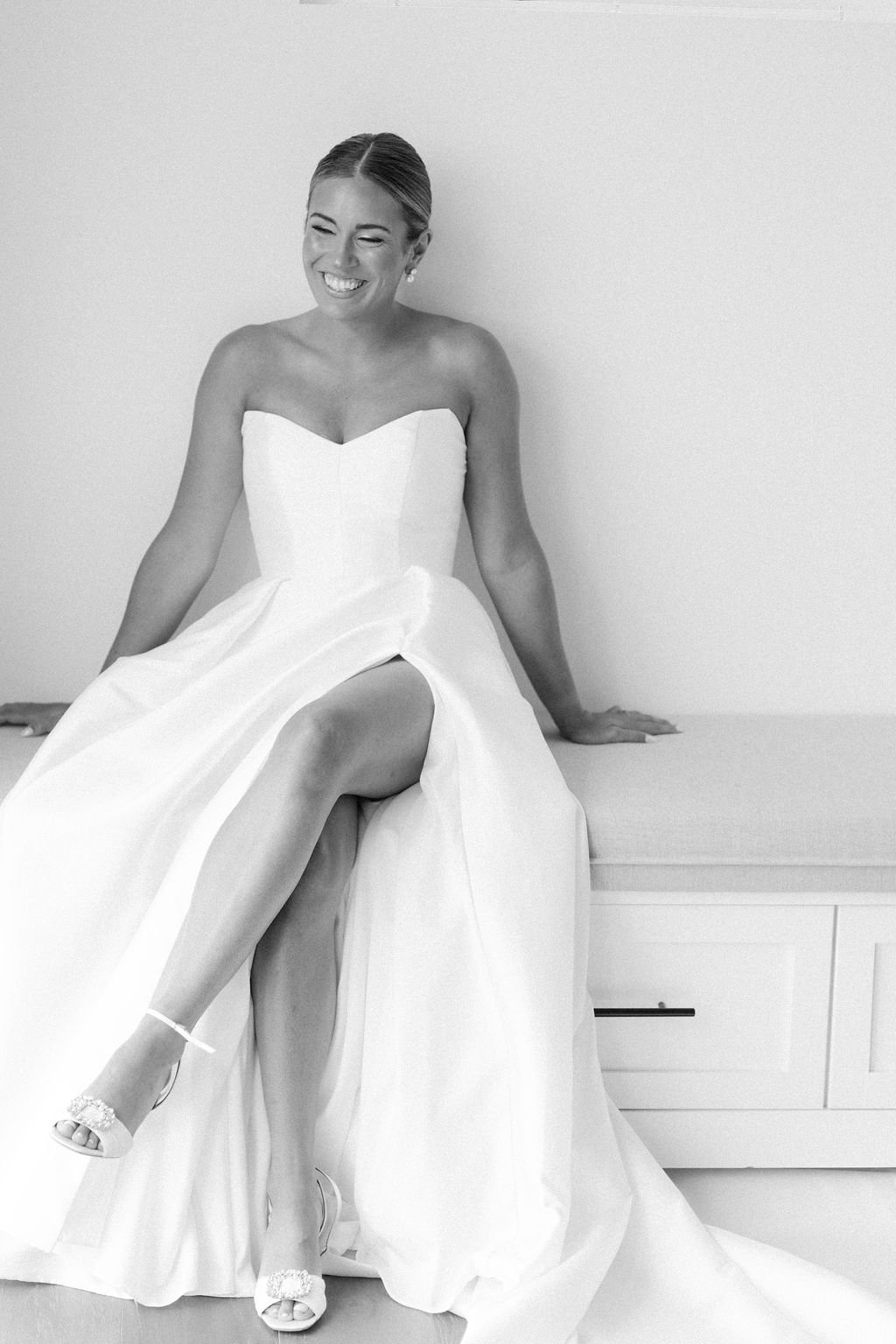 Woman in a wedding dress sitting on a table, smiling with eyes closed.