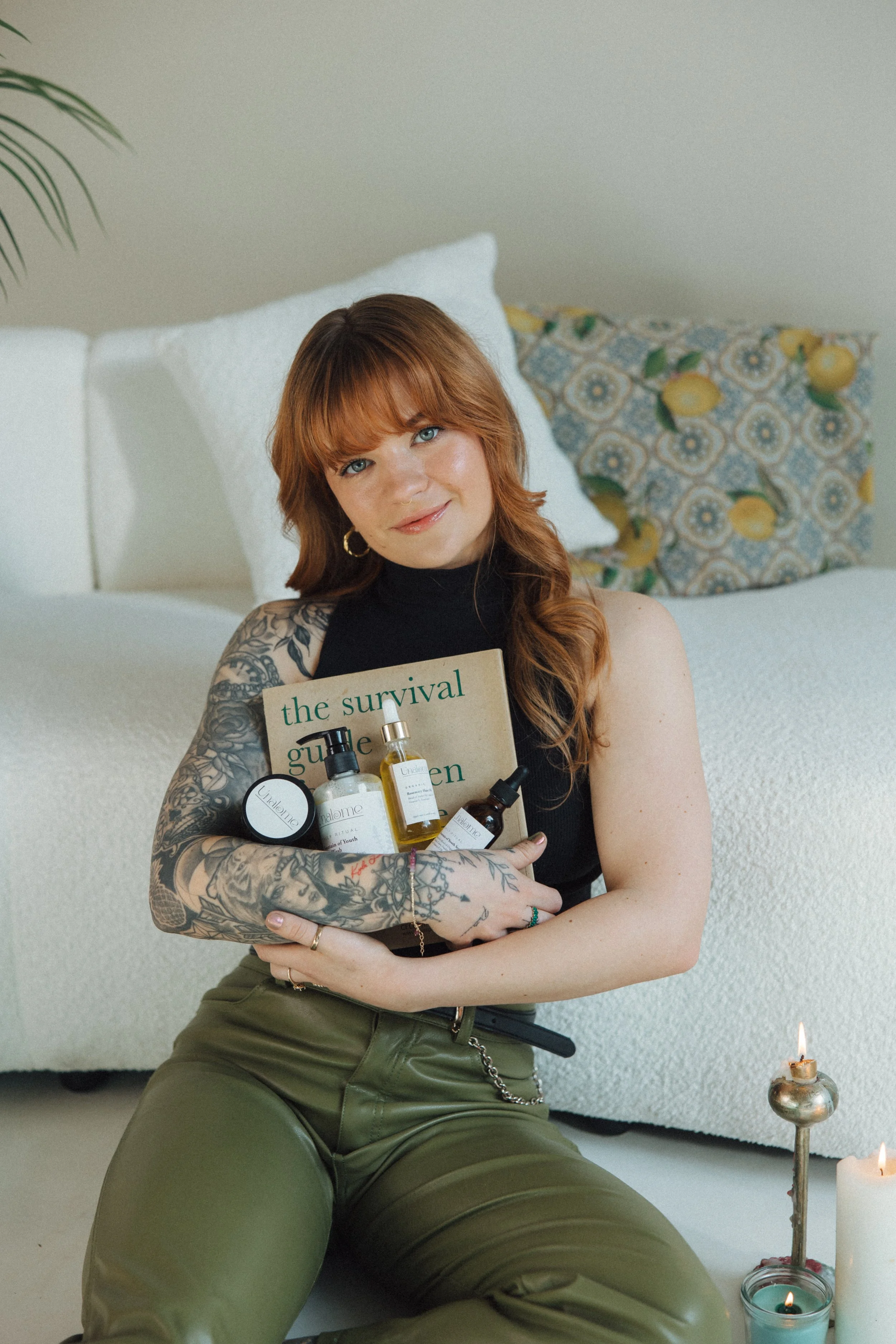 A woman with red hair and tattoos on her left arm is sitting on the floor holding a bottle of skincare product and a magazine. She is wearing a black sleeveless top and olive green pants. There are candles and a candle holder nearby on the floor.