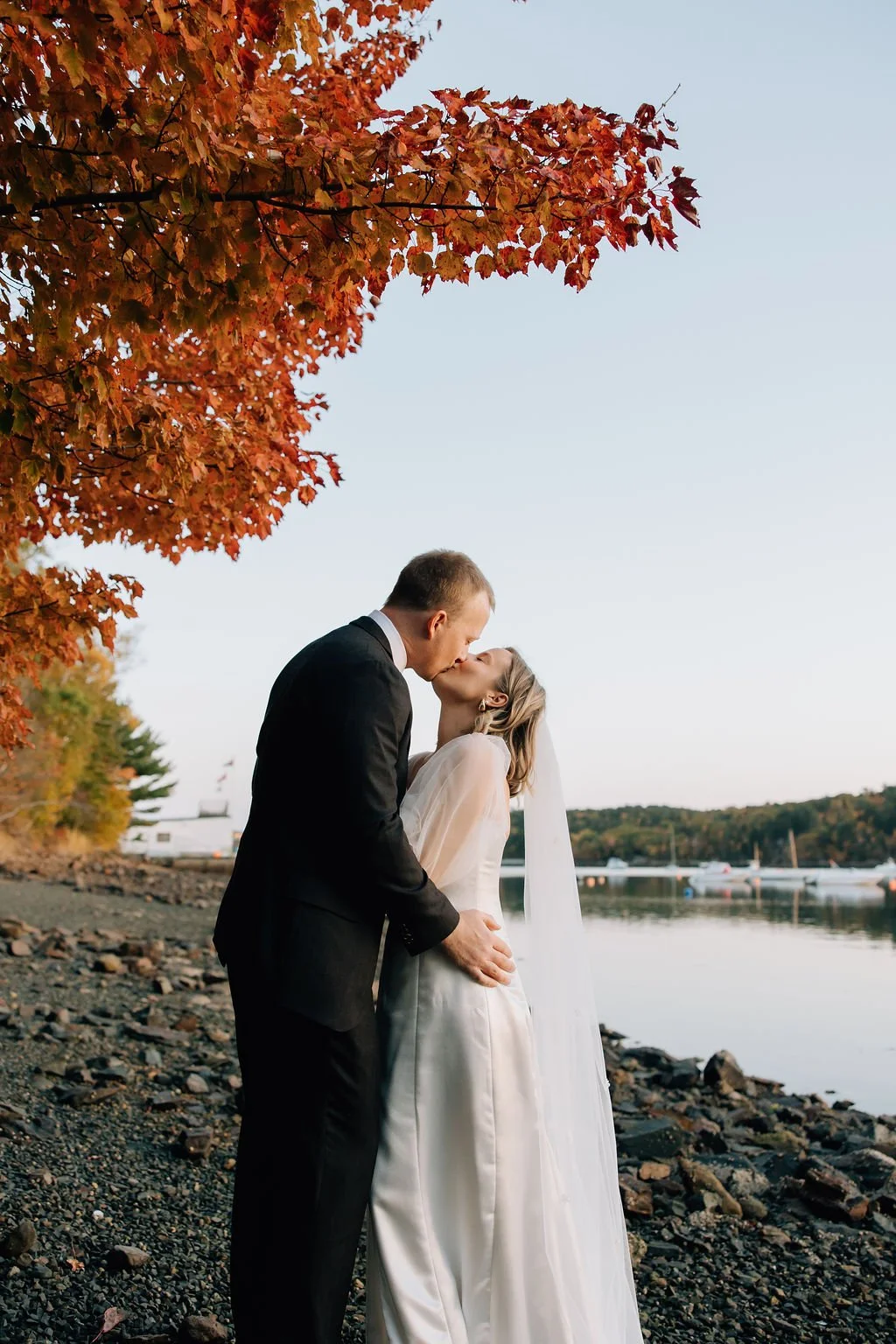 A bride and groom share a kiss on a rocky riverside during fall, with red and orange autumn leaves overhead and boats on the calm water in the background.