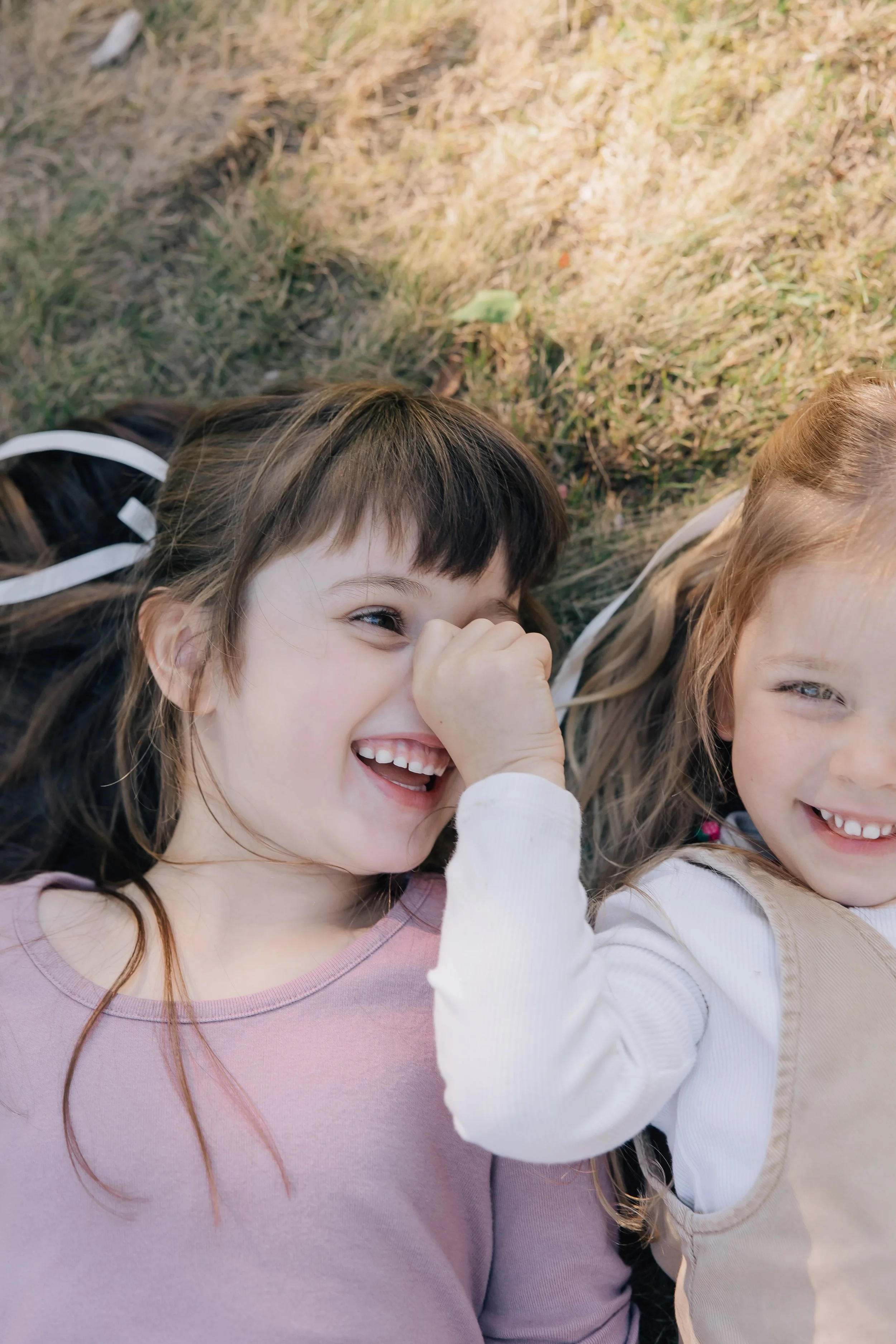 Two young girls lying on the grass outdoors, laughing and smiling, with one girl touching her nose.