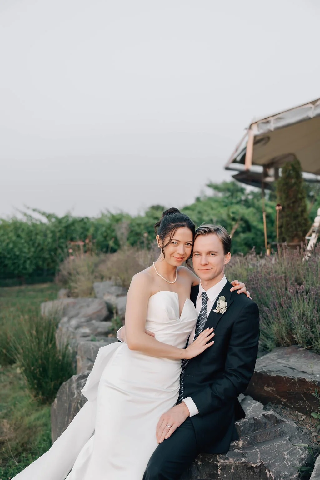A newlywed couple is sitting on rocks outdoors with greenery and purple flowers in the background, smiling at the camera. The bride is wearing a strapless white wedding dress and pearl necklace, and the groom is in a black suit with a white shirt and