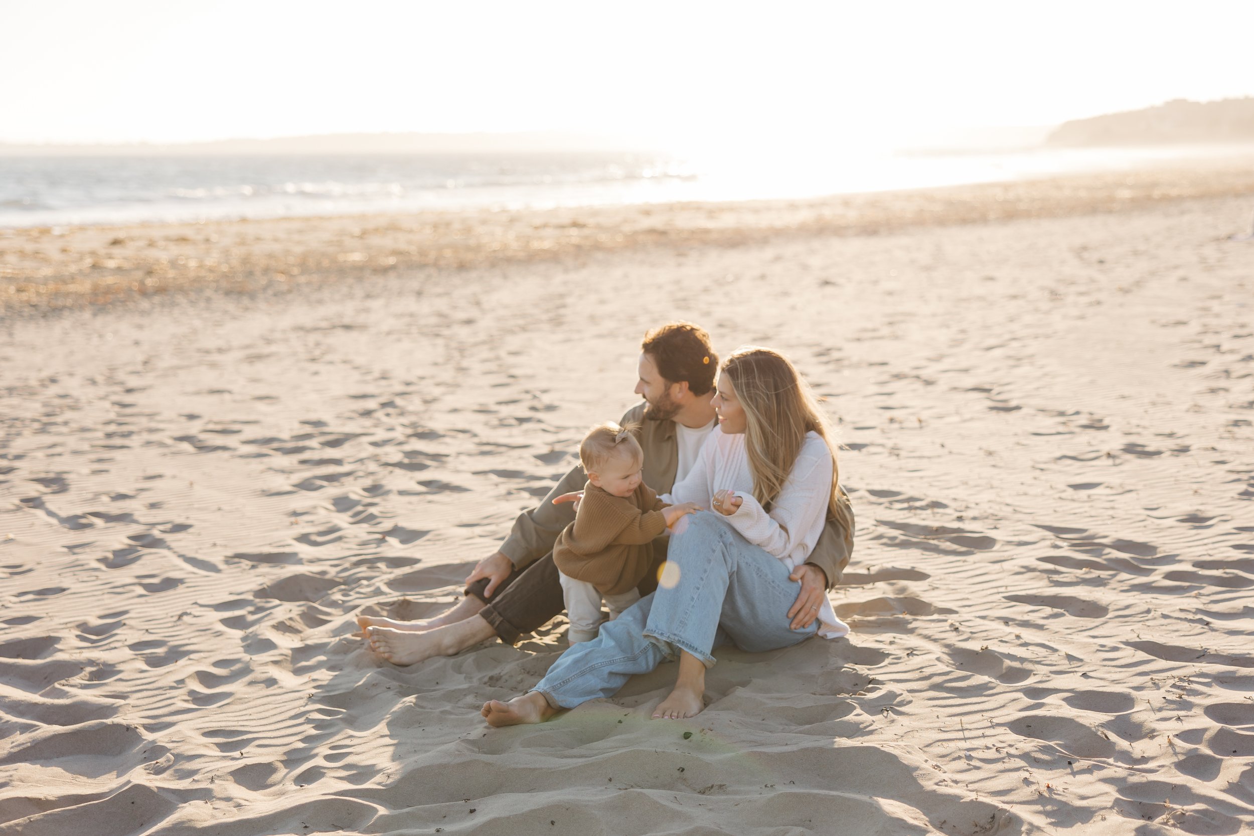 A family of three sitting on the sandy beach, smiling and spending quality time together during sunset, with the ocean and distant land in the background.