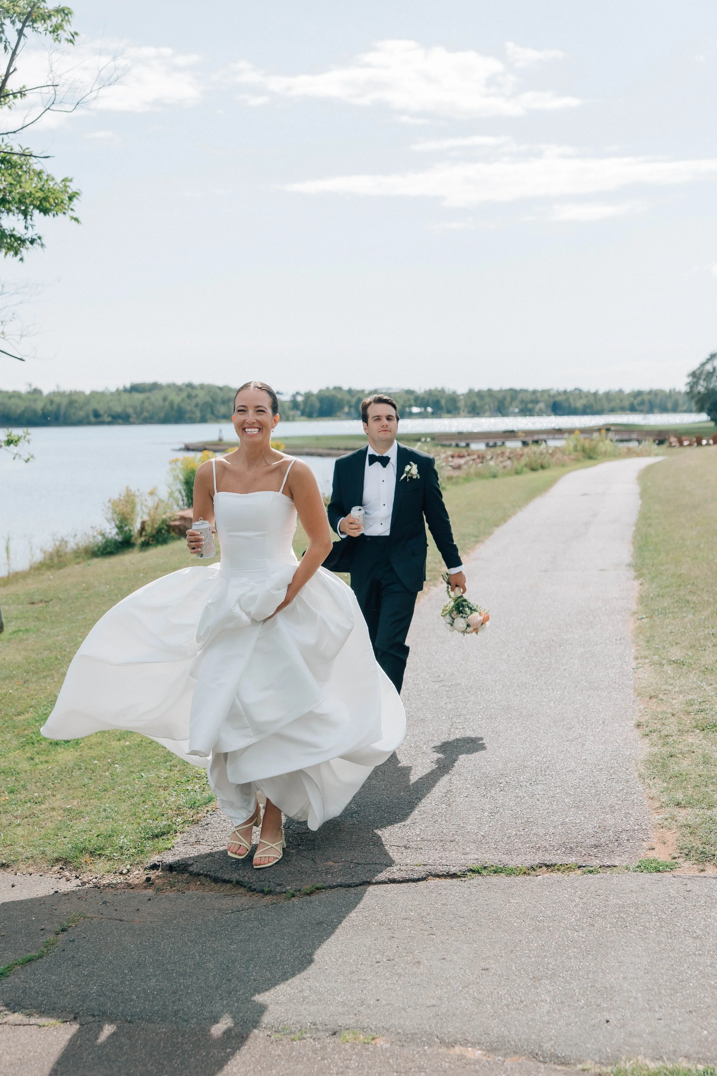A bride in a white wedding gown walking outdoors along a pathway next to a lake, holding a white bouquet, with a groom in a tuxedo walking behind her, holding a bouquet of flowers, sunny weather, trees, and water in the background.