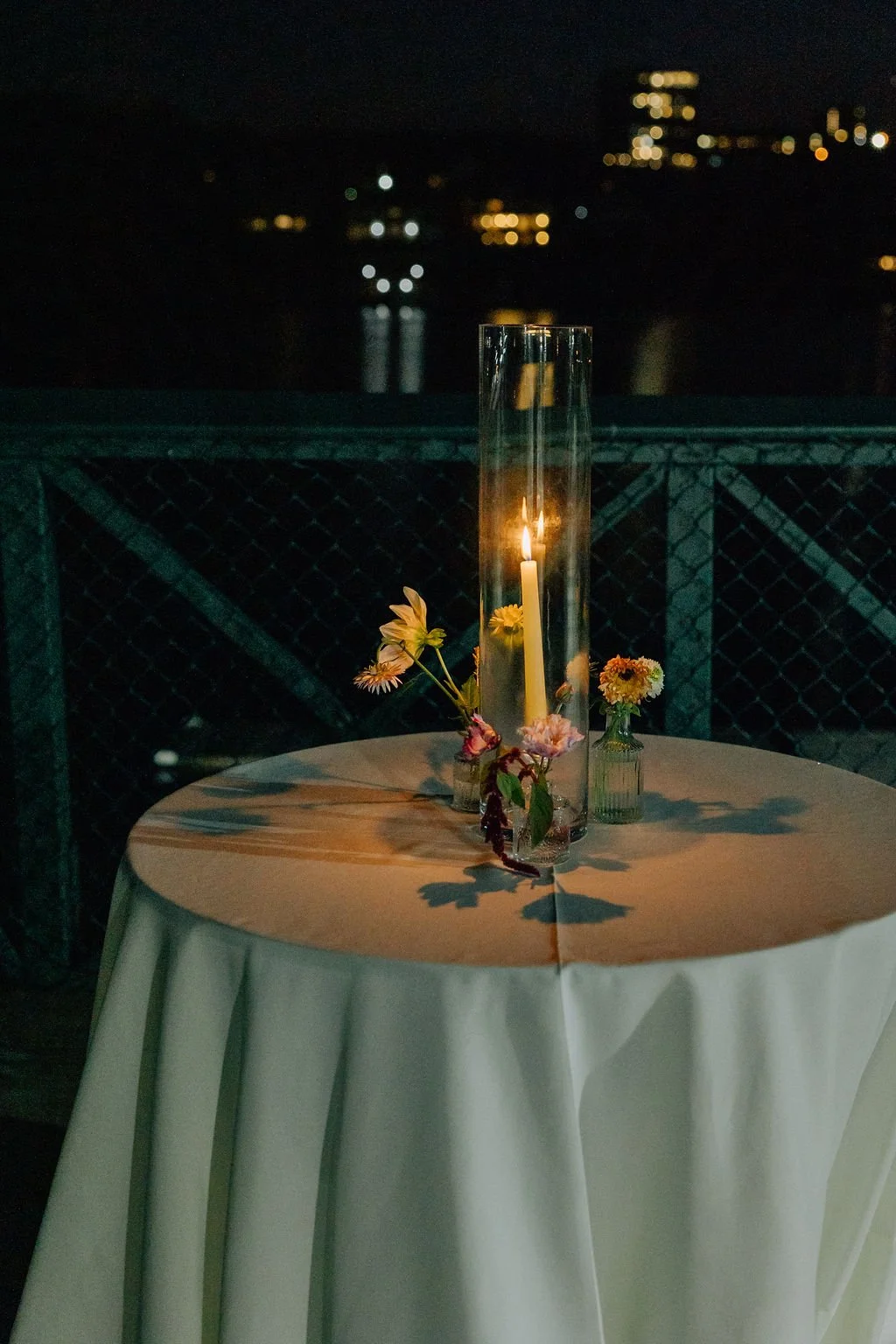 A round table with a white tablecloth, a tall glass candle holder with a lit candle, and small vases with flowers, set against a dark night background with city lights in the distance.
