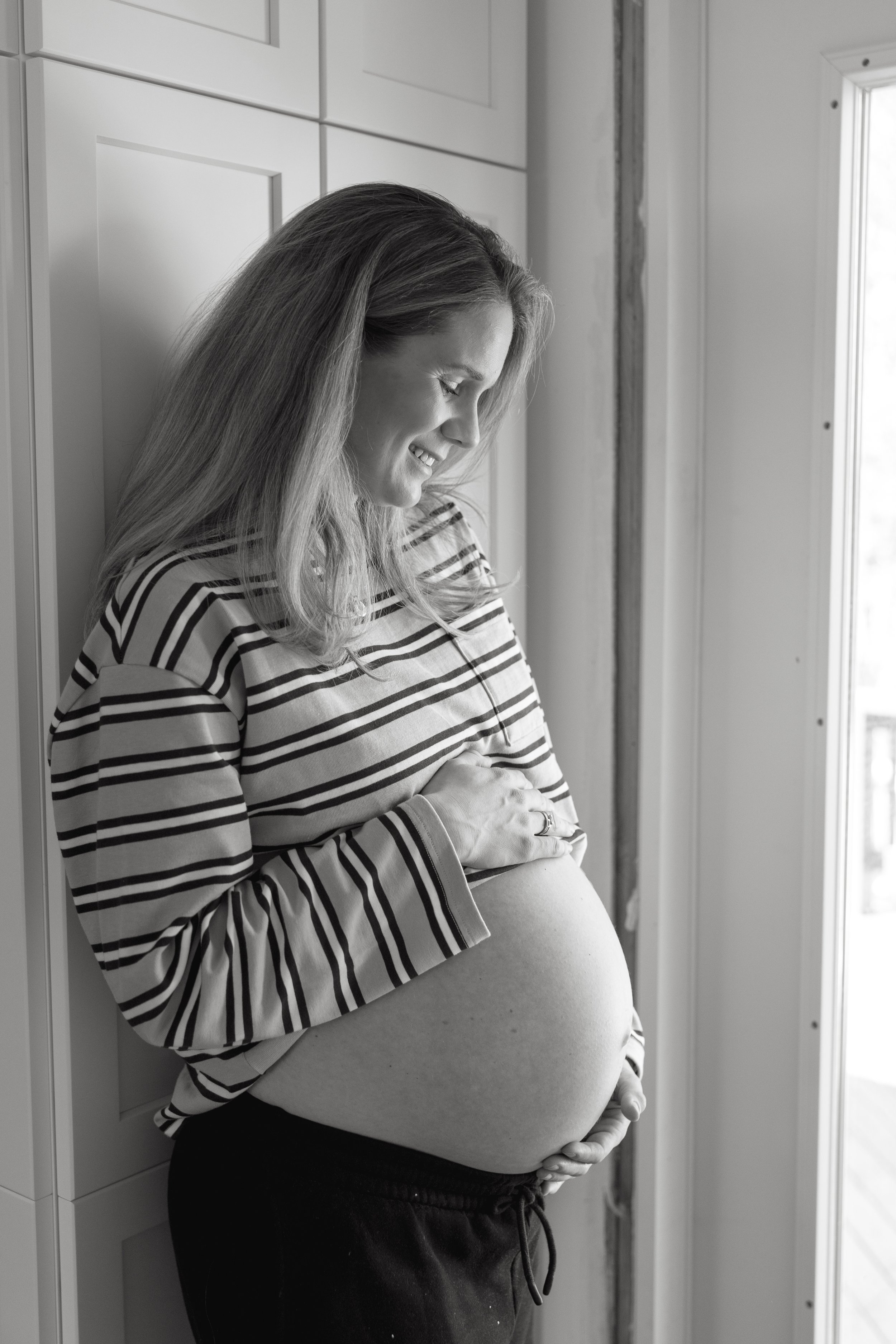 Black and white photo of a pregnant woman smiling while standing by a window, with one hand on her belly and the other hand supporting her lower back.
