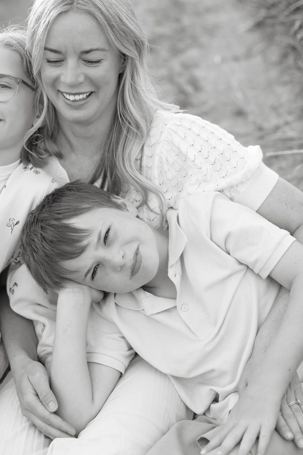 A woman and two boys lying together outdoors, smiling and relaxing with one boy resting his head on his hand, all in a black and white photo.