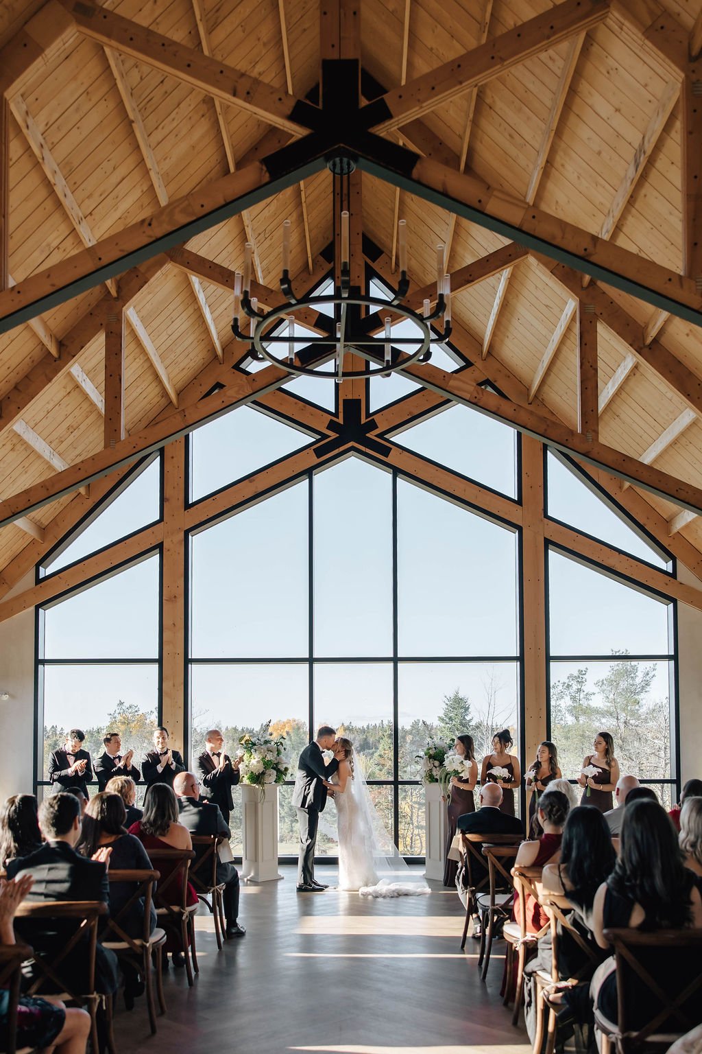 A wedding ceremony with large glass windows, capturing the bride and groom kissing at the altar, surrounded by wedding party and guests.