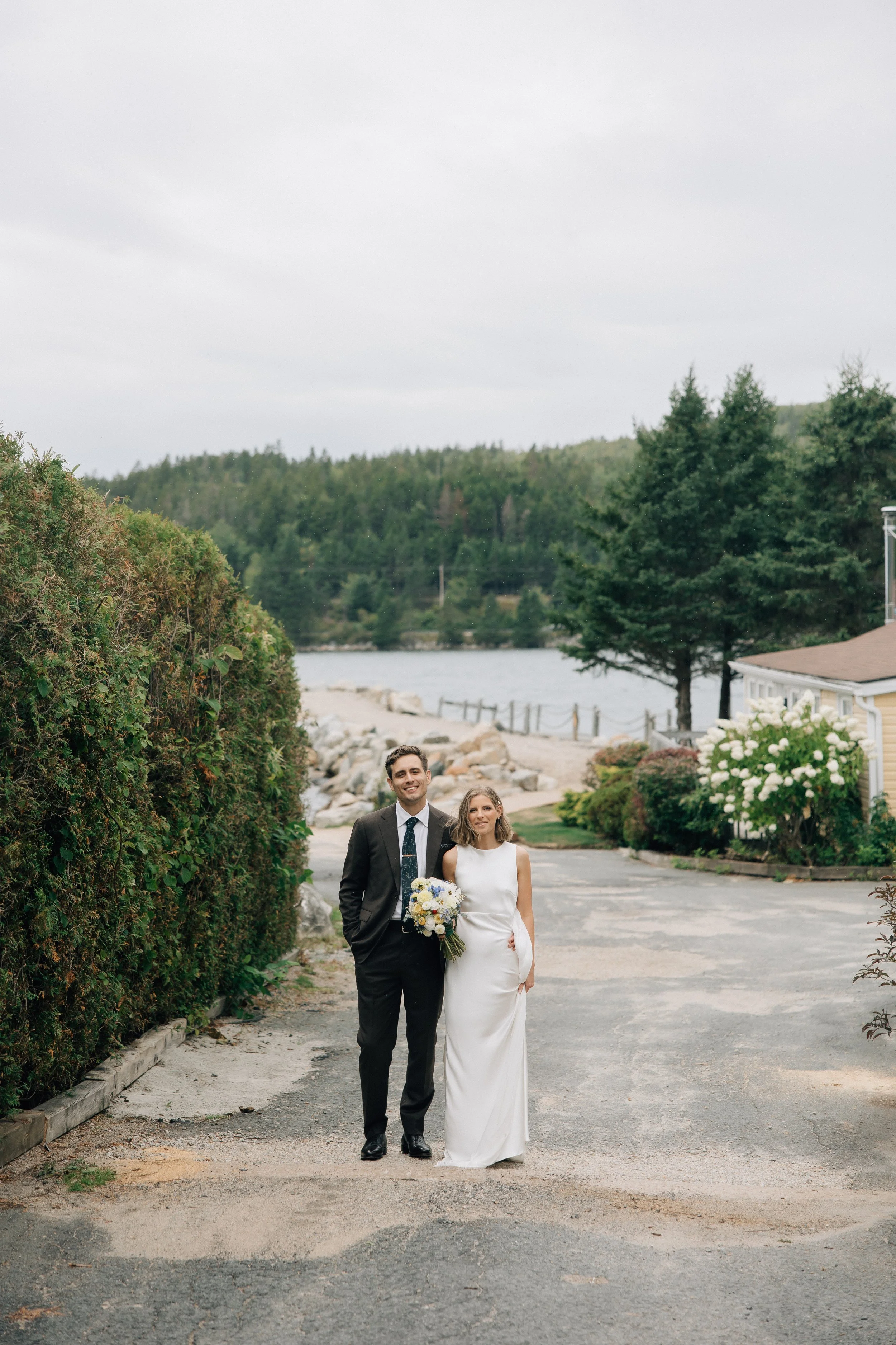 A newlywed couple walking outdoors on a gravel path near water, with the groom in a black suit and the bride in a white wedding dress holding a bouquet, surrounded by greenery and trees on a cloudy day.