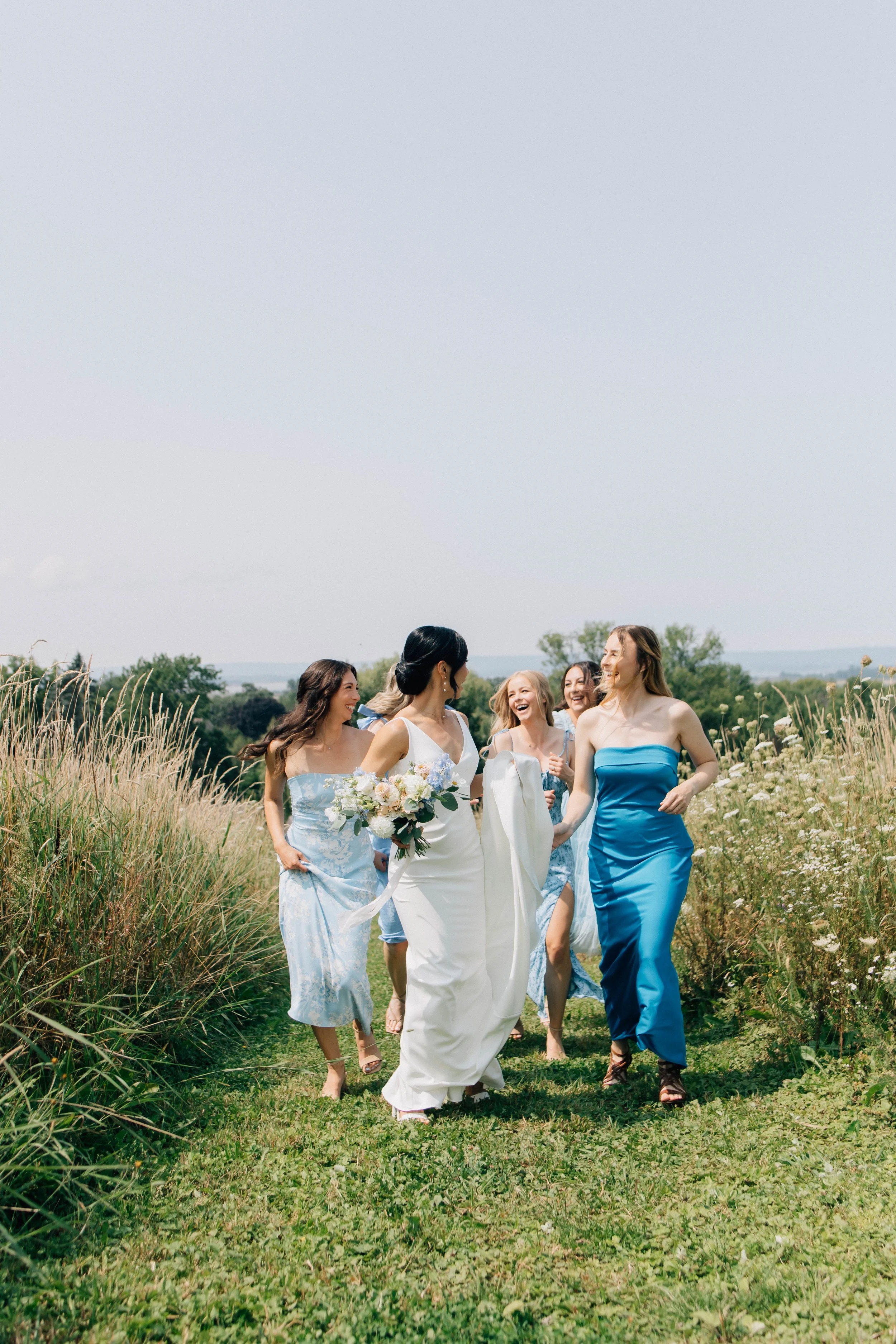 A group of women walking outdoors in a grassy field, dressed in formal and semi-formal attire, smiling and enjoying each other's company.
