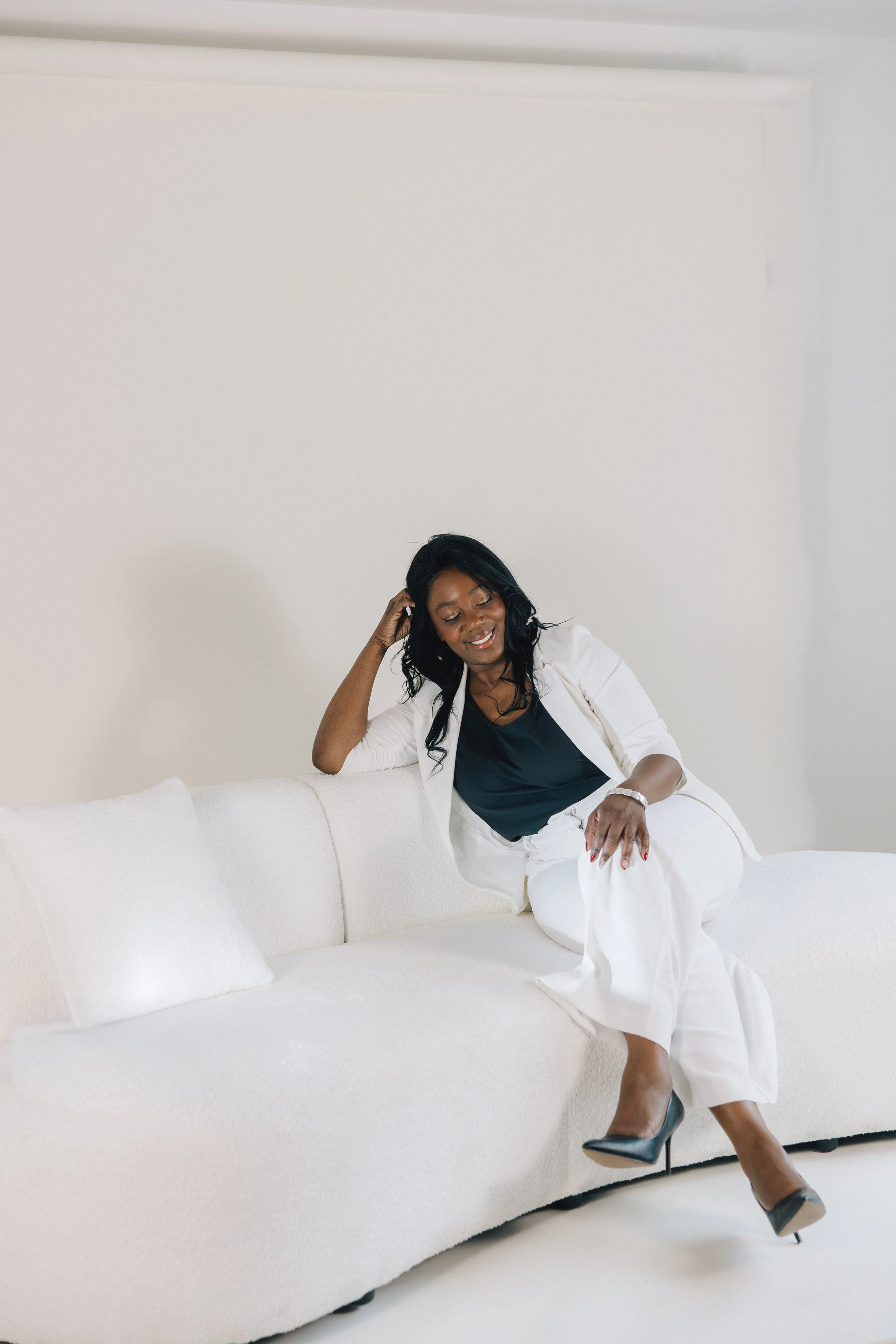 A woman with black hair, wearing a white blazer and white pants, is sitting on a white couch with white pillows, smiling and relaxing with one hand on her head and the other on her knee, in a minimalistic white room.