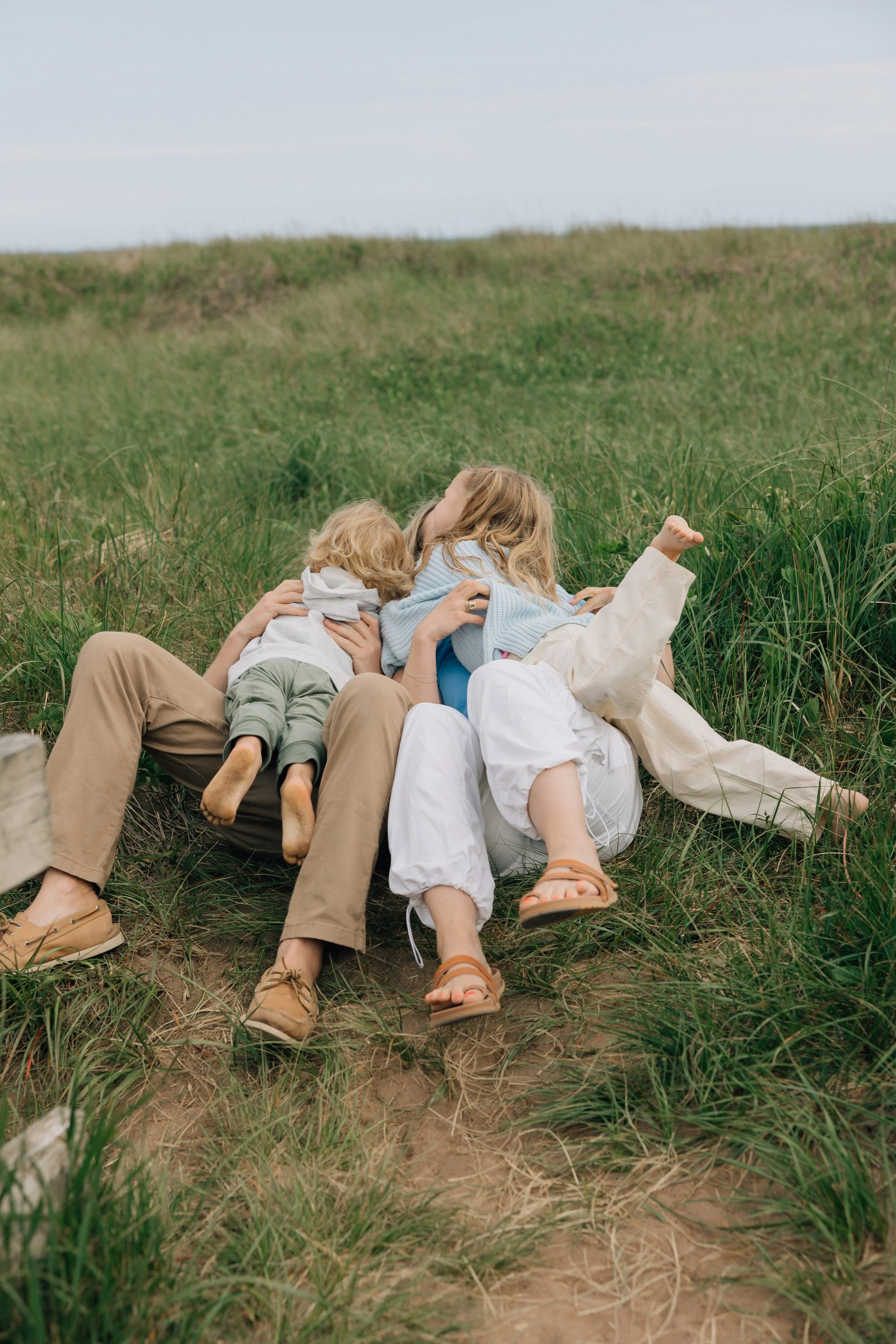 A group of three people, likely a family, lying on the grass in a field with tall green grass, embracing each other and seemingly sharing a moment of affection. The sky is partly cloudy.