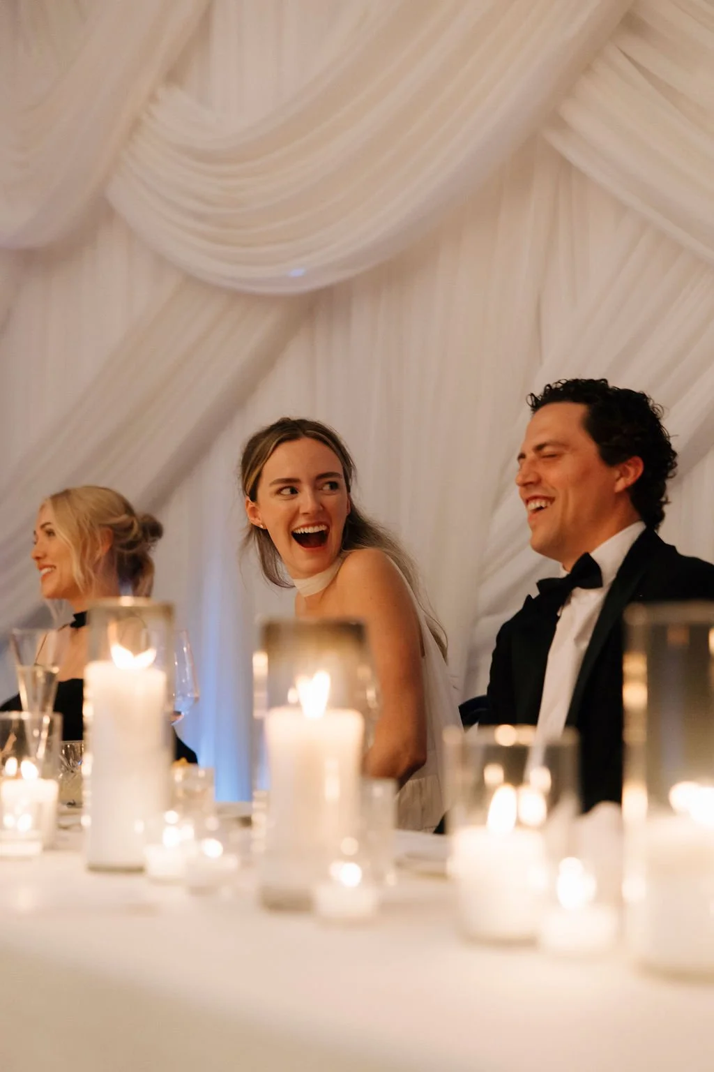 People sitting at a table during a wedding reception, smiling and laughing, with candles on the table and draped fabric on the ceiling.