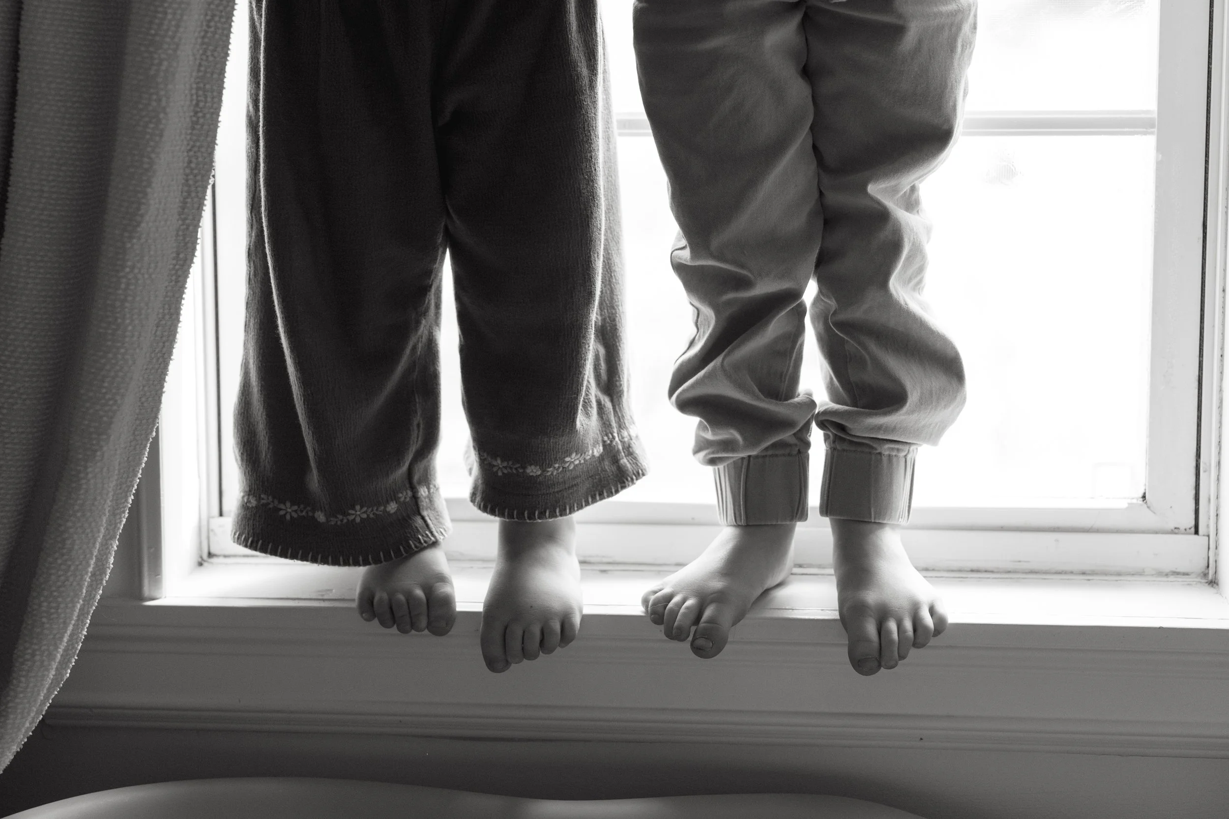 Children's legs and feet standing on a windowsill. One child wearing dark pants with a floral pattern at the bottom, and the other child wearing light-colored pants with cuffs. Both are barefoot, and the background shows bright light coming through t