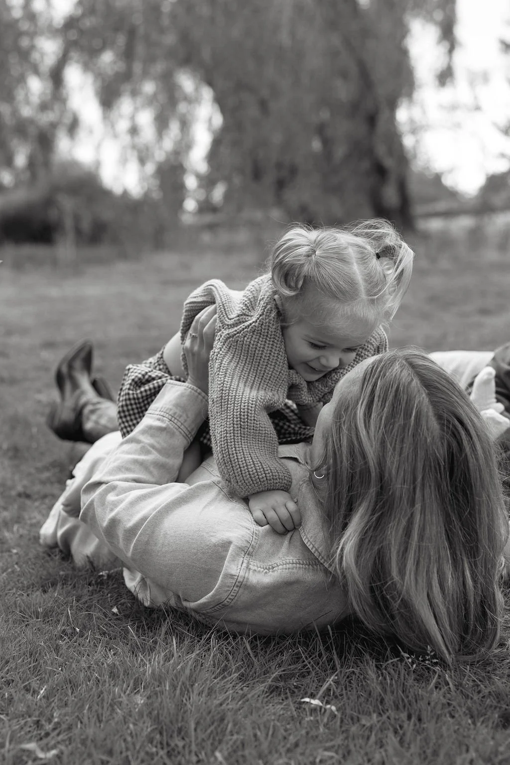 A child and an adult playing on the grass outdoors, the child lying on top of the adult with their faces close together and smiling.