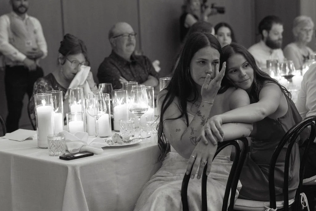 Two young women sitting at a table during a formal event, one embracing the other, with candles and glasses on the table in a dimly lit room.
