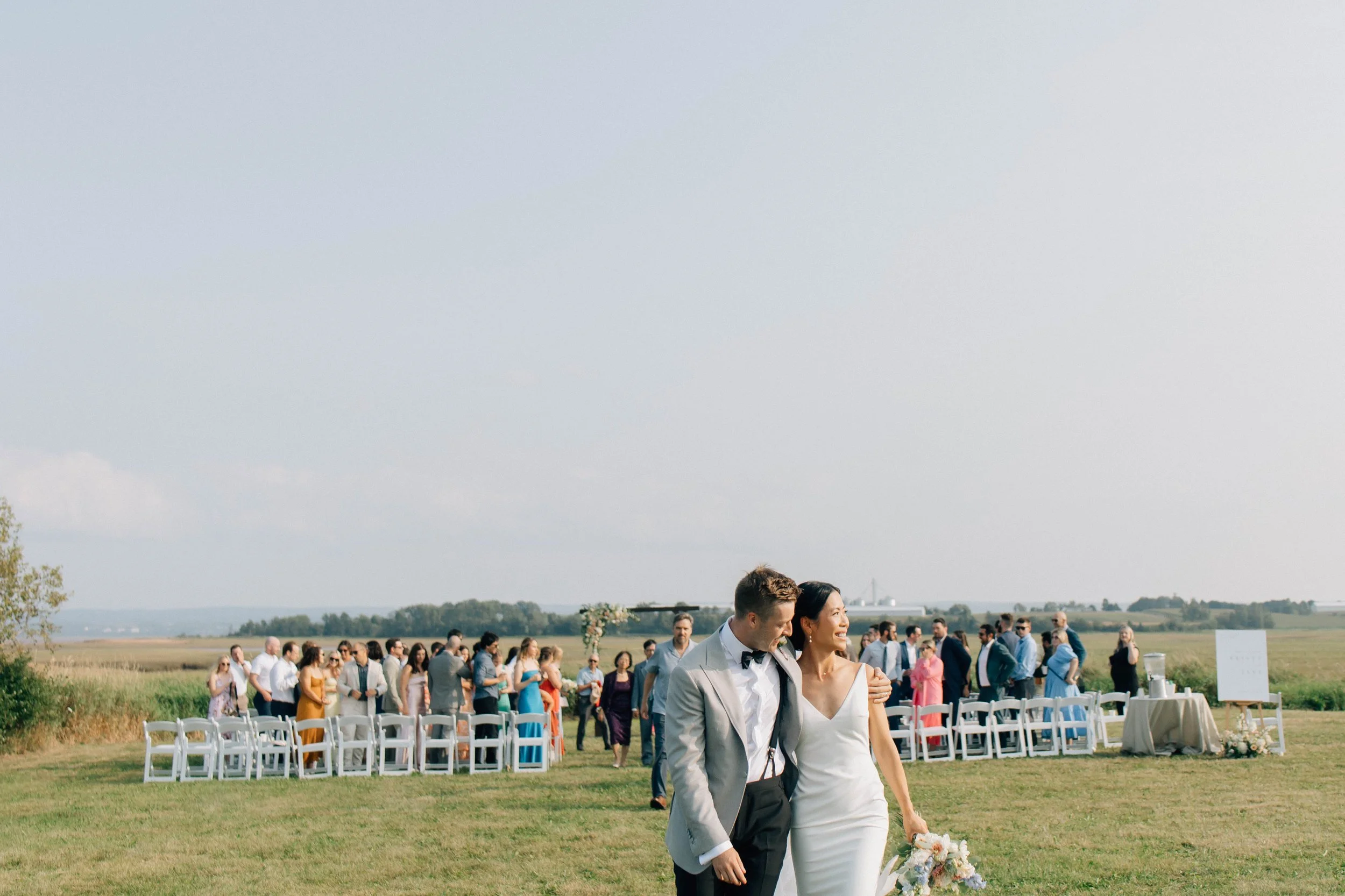 A bride and groom walking hand in hand at an outdoor wedding ceremony on a sunny day, with guests seated in white chairs and standing in the background.