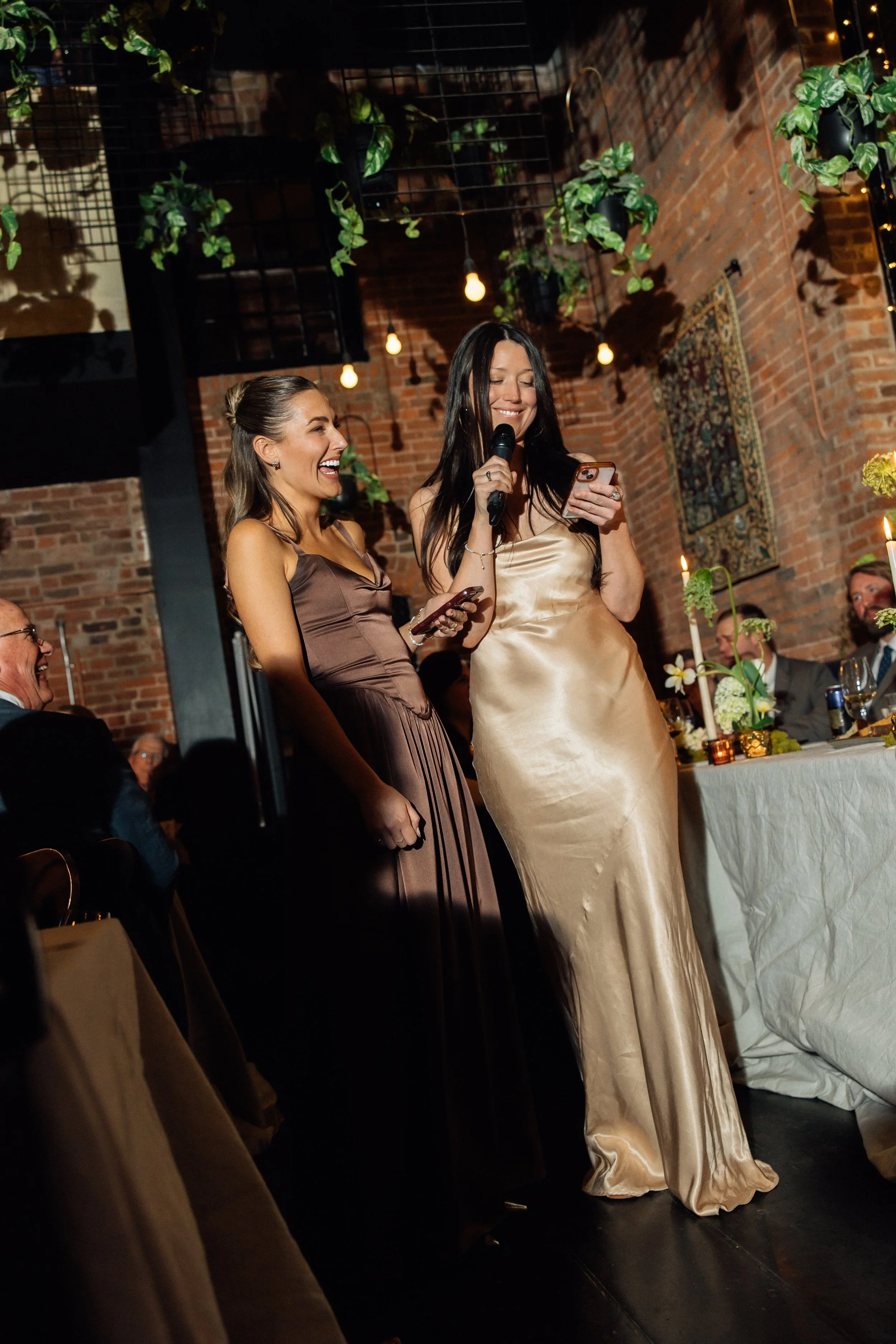 Two women in elegant dresses are standing together at a celebration event, smiling and looking down at their phones, with a festive atmosphere including candles and decorative plants in a warmly lit brick venue.