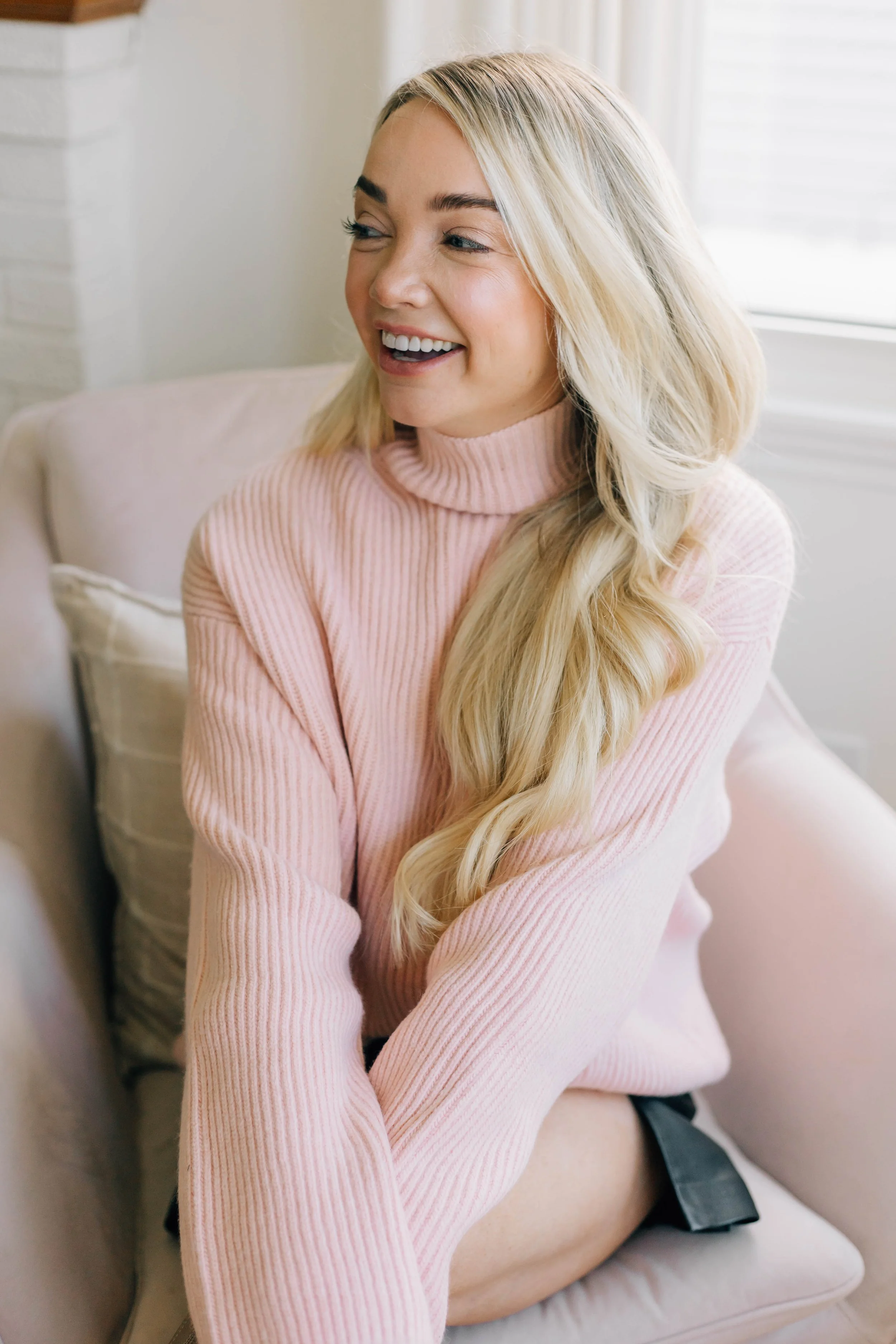 A woman with long blonde hair smiling while sitting on a beige couch, wearing a pink turtleneck sweater.