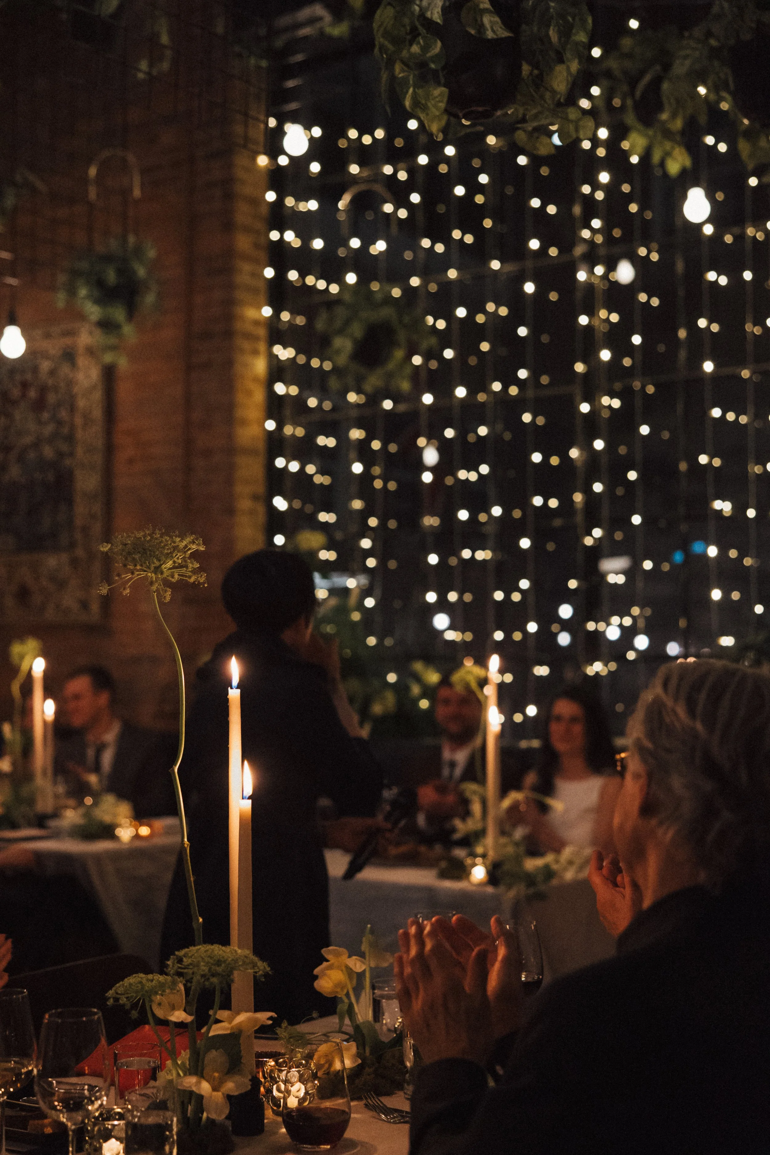 People gathered around a dinner table decorated with flowers and candles, at an outdoor evening event with string lights and a grid of small lights in the background.