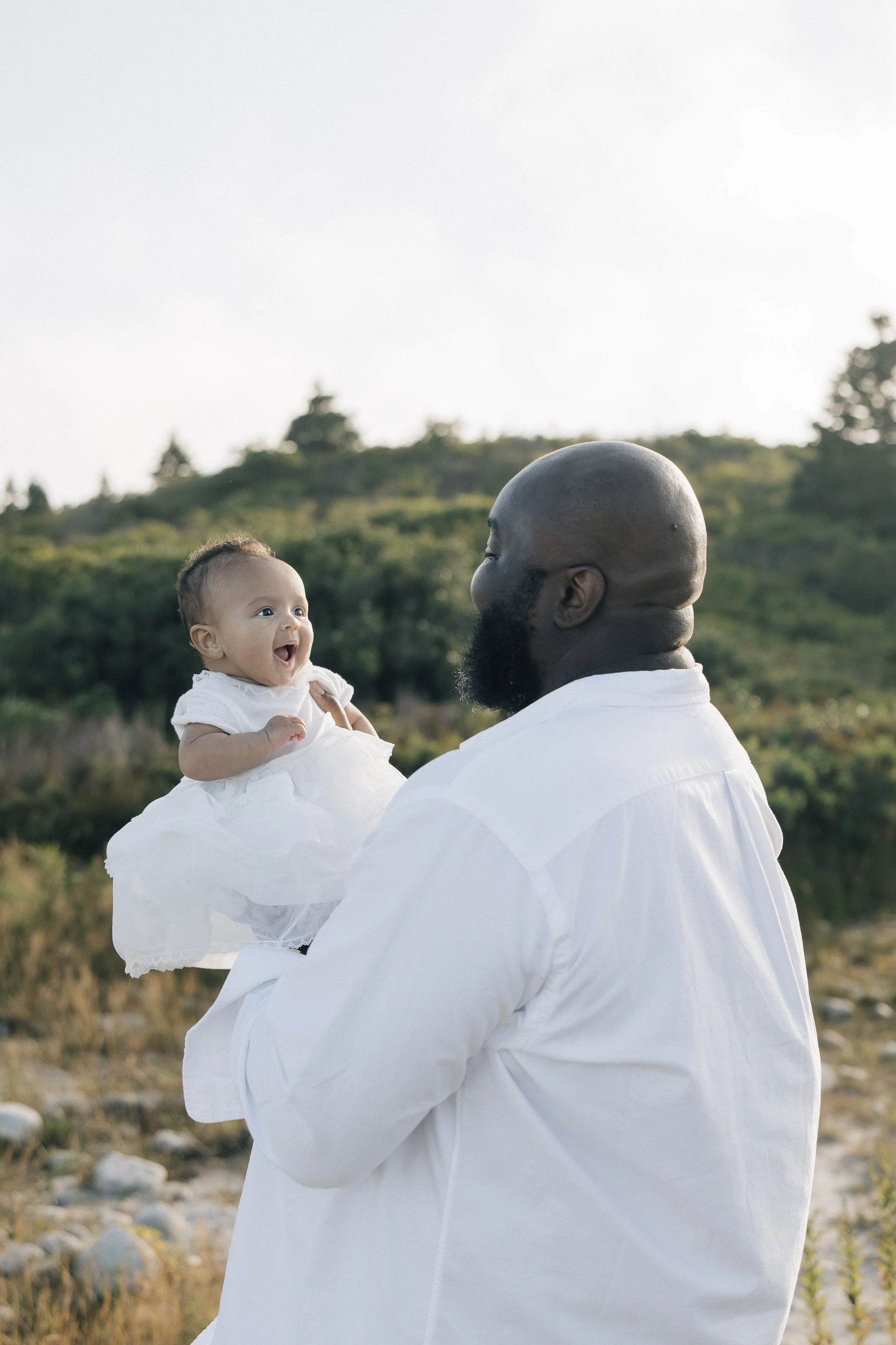 An African American man with a beard holding a smiling baby girl in an outdoor natural setting with greenery and trees in the background.