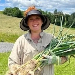 Photo of a woman standing in a field wearing a hat. She looks at the camera smiling. She is holding a large bunch of onions
