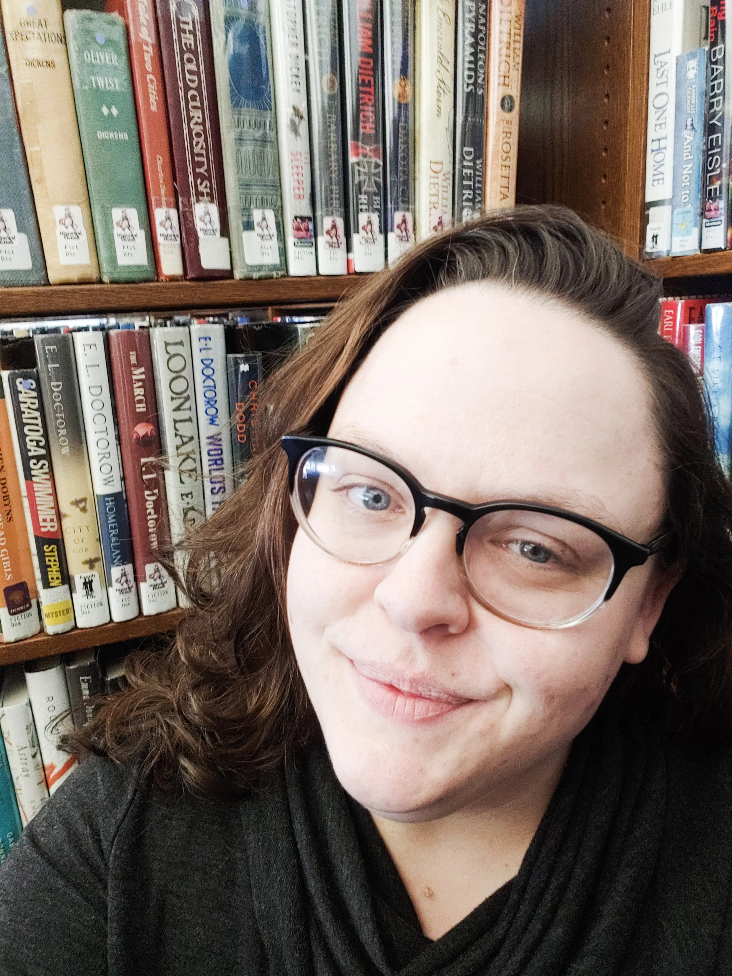 Selfie of a woman in front of a shelf of books. She has brown hair and wears glasses