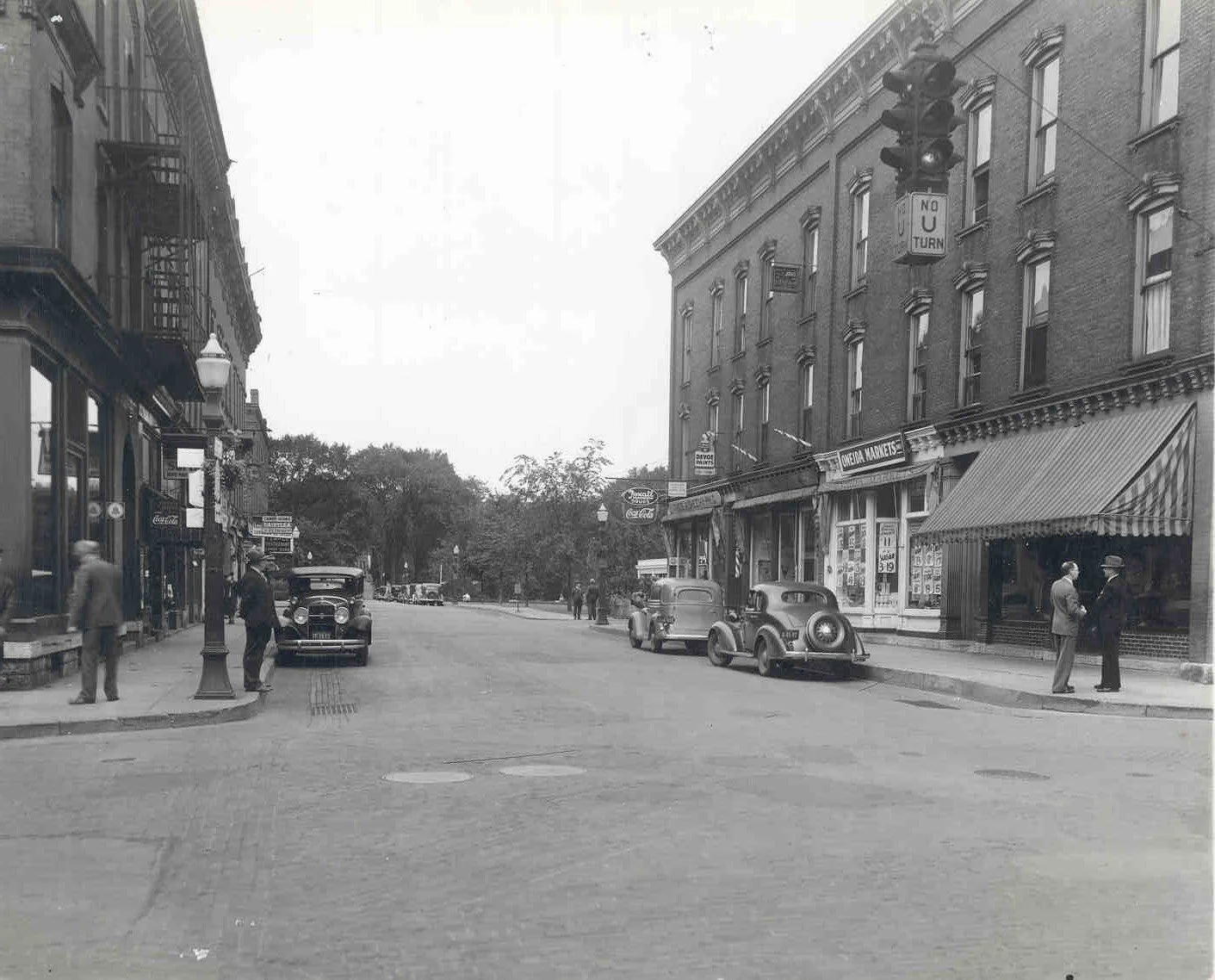 Black and white photo of a street with brick buildings and trees in the background. 