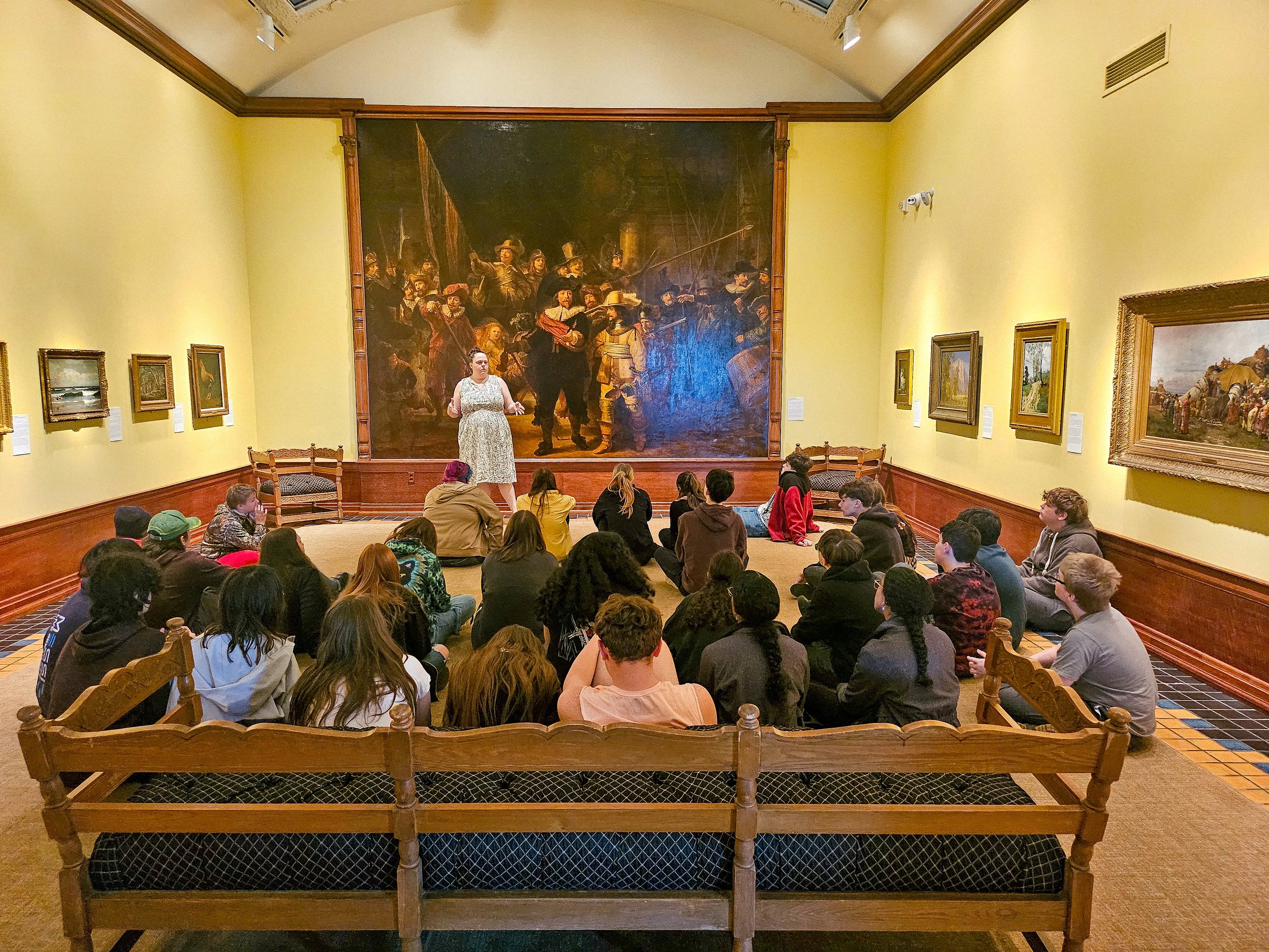 This image features Curator of Education and Public Engagement Mary Alexander presenting information about our Night Watch gallery to a group of 30 students; Their backs are turned toward the camera and they are seated in lecture format.