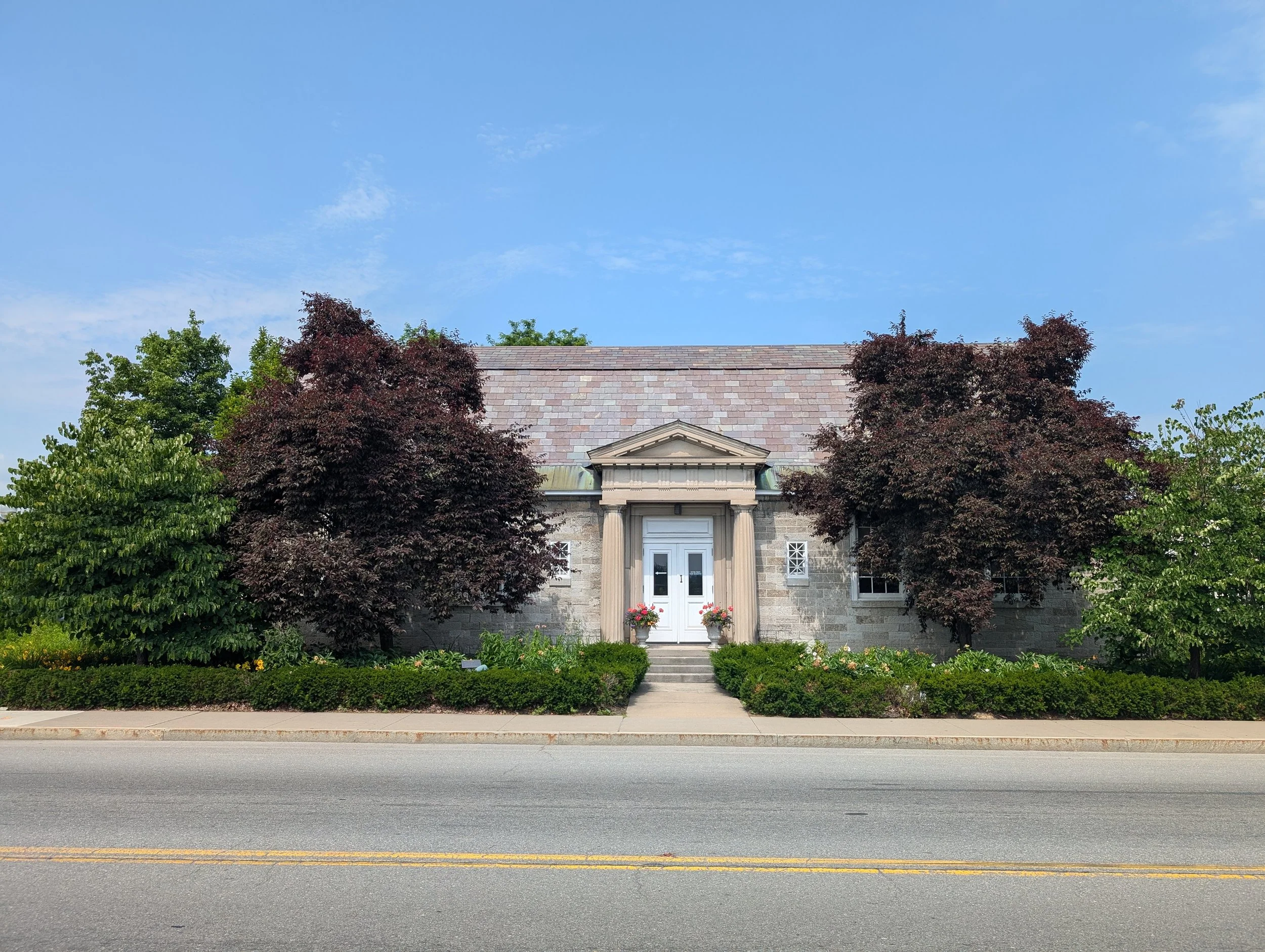 Picture of a stone building with a Greek style entryway. There are purple leafed trees out front to the right and left of the central door.