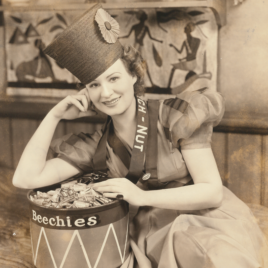 Black and white photo of a woman wearing a majorette uniform. She leans on a drum filled with samples of Beech-Nut gum. 