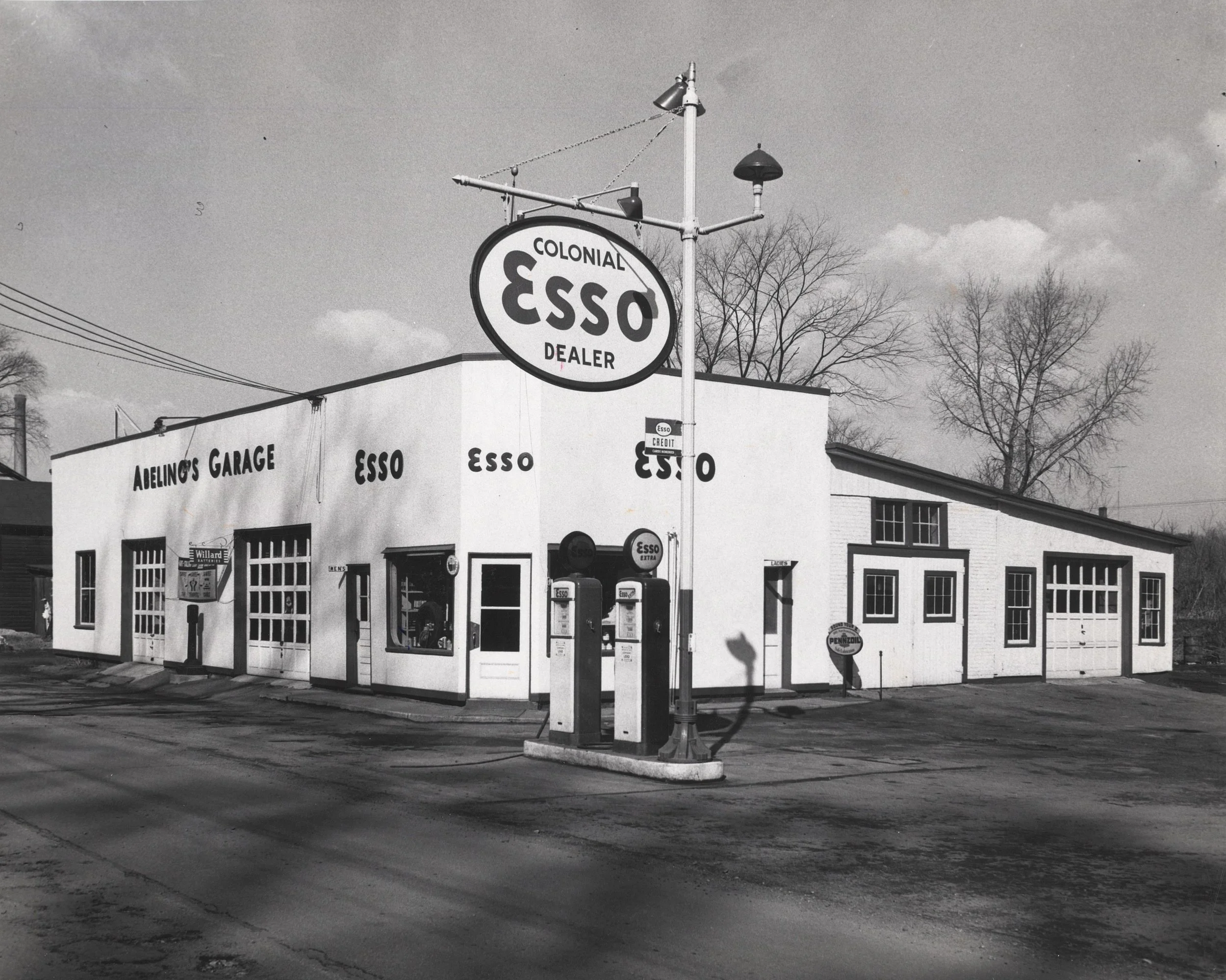 Black and white photograph of an Esso Gas Station. The station is a large white building with garage bays and two gas pumps out front. 