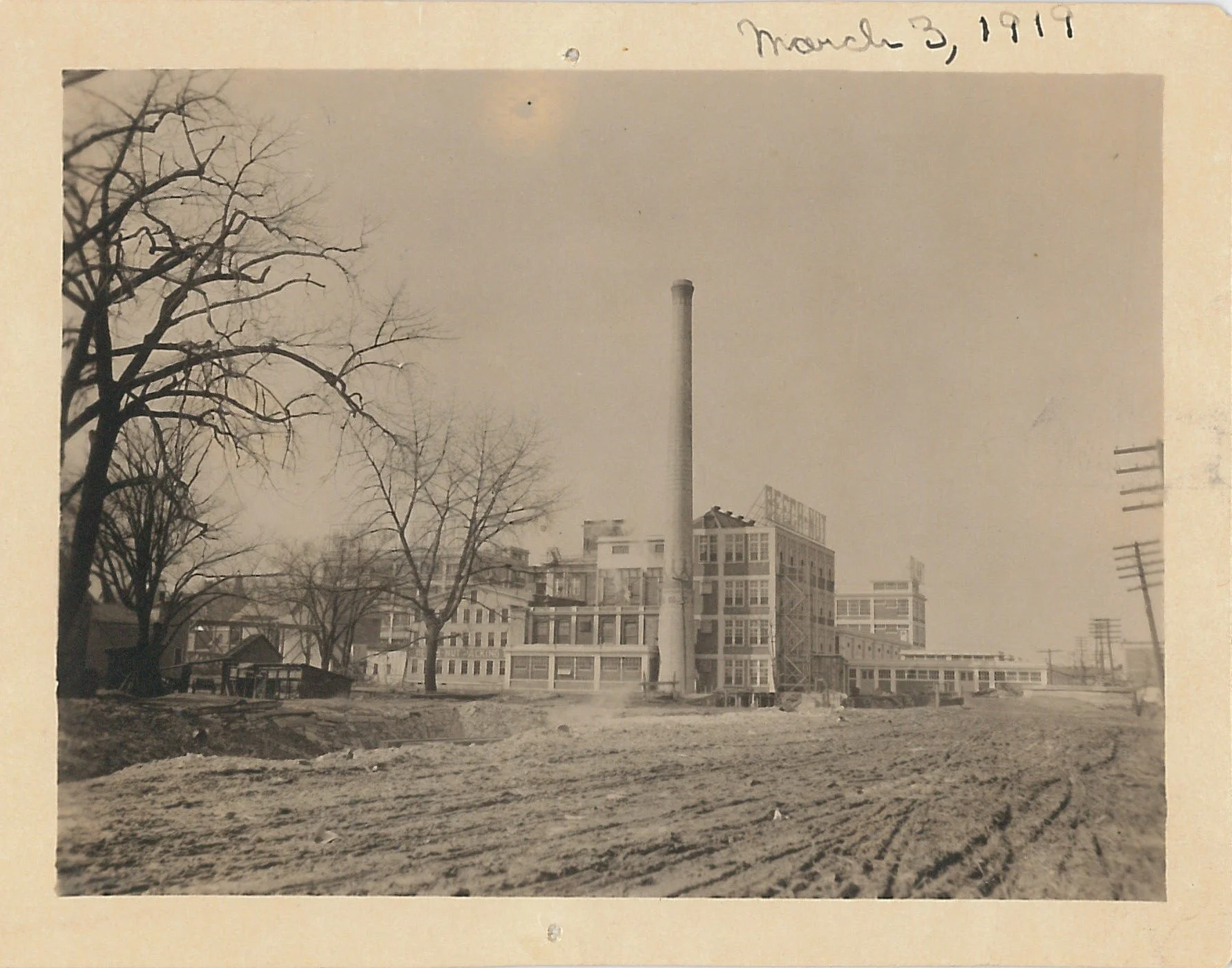 Black and white photo of a large dirt road like area, behind there is a smoke stack and factory.