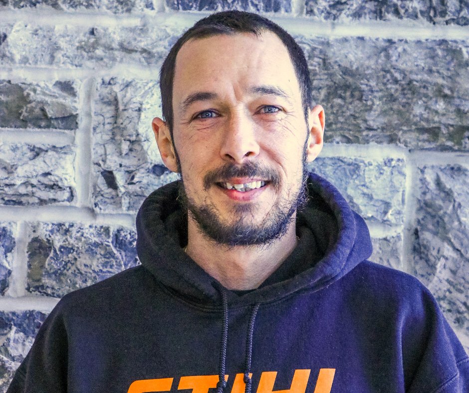 Man standing in front of a stone wall. He looks at the camera and smiles he is wearing a blue hoodie with orange writing on it