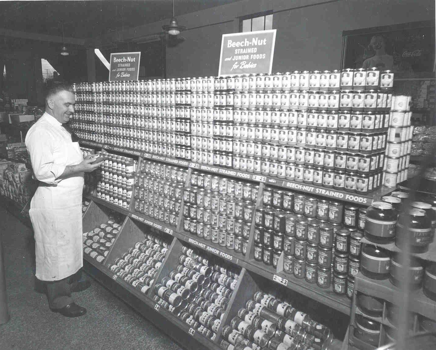Black and white photo of a man in a white apron standing in front of a large display of Beech-Nut Junior Foods. 