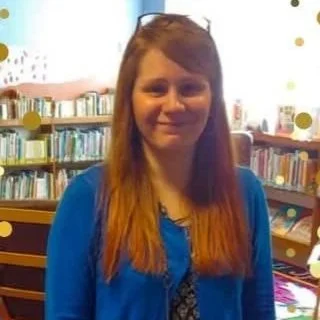Photo of a woman in a blue sweater looking at the camera. She has long medium brown hair and has a pair of glasses on her head..