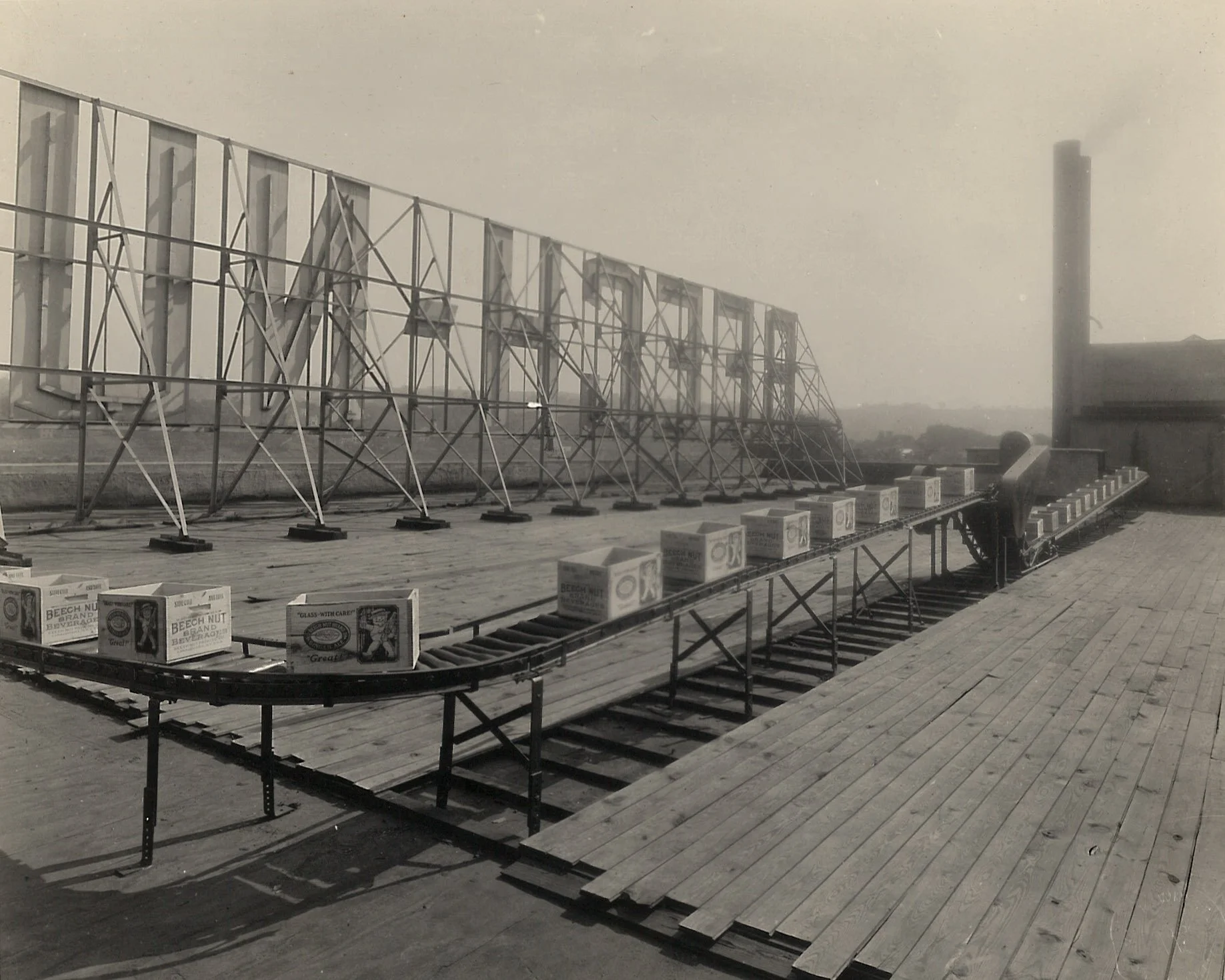 Black and white photo of the roof of a packing company. A conveyor belt moves boxes of ginger ale over the roof. There is the back of a large sign that reads: Beech-Nut