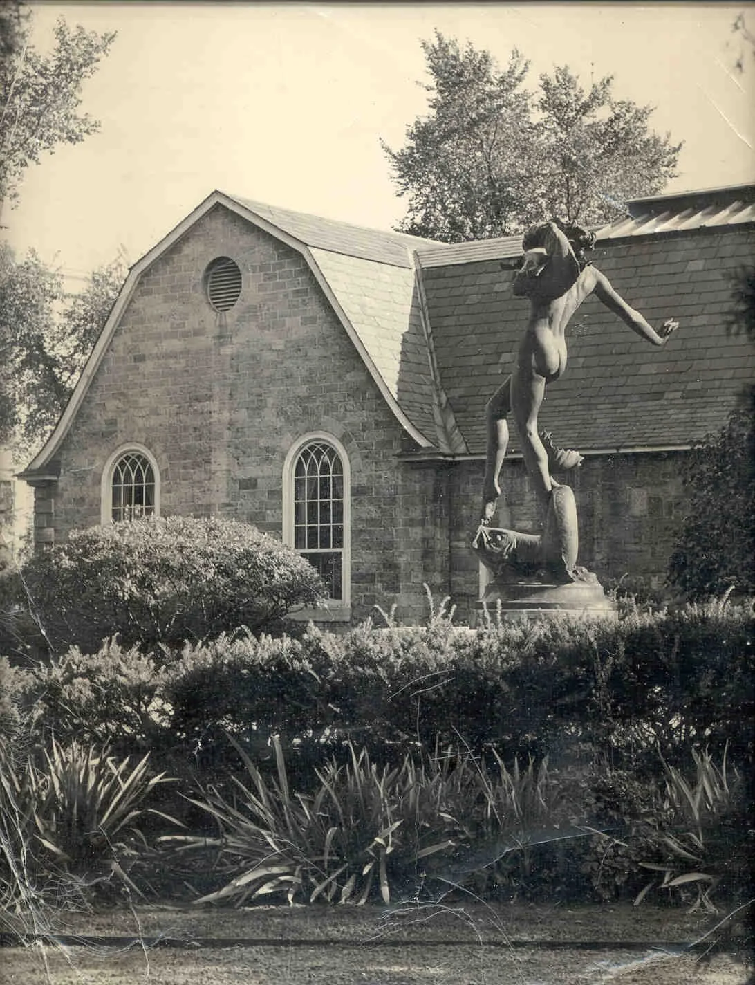 Black and white photo of a garden with a statue of a naked woman standing on a fish. There is a stone building in the background