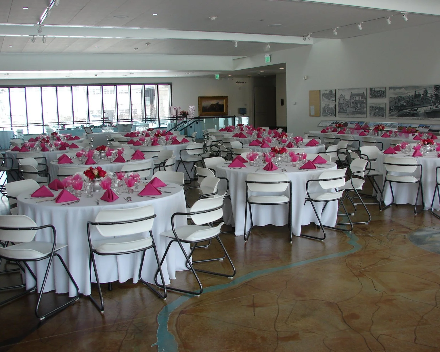 This is a photo of the Arkell Museum and Canajoharie Library Great Hall. It is currently staged for a party; There is approximately 8 large round tables with chairs at each table. They are dressed with white linens, glassware, and pink napkins.
