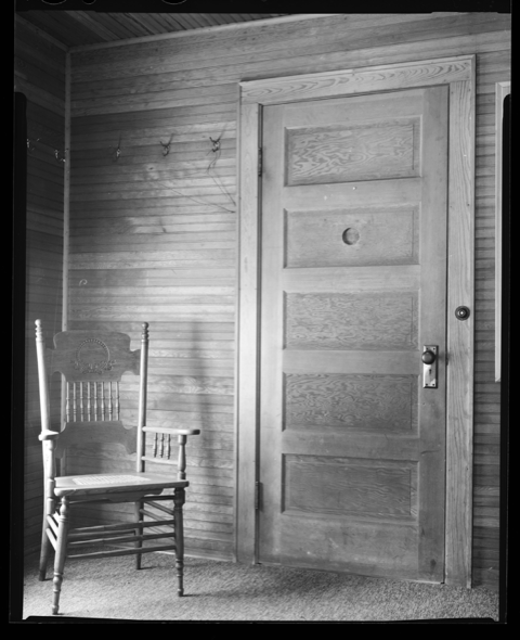 Black and white photo of a wooden door with a peep hole in it. There is a wooden chair next to the door. 
