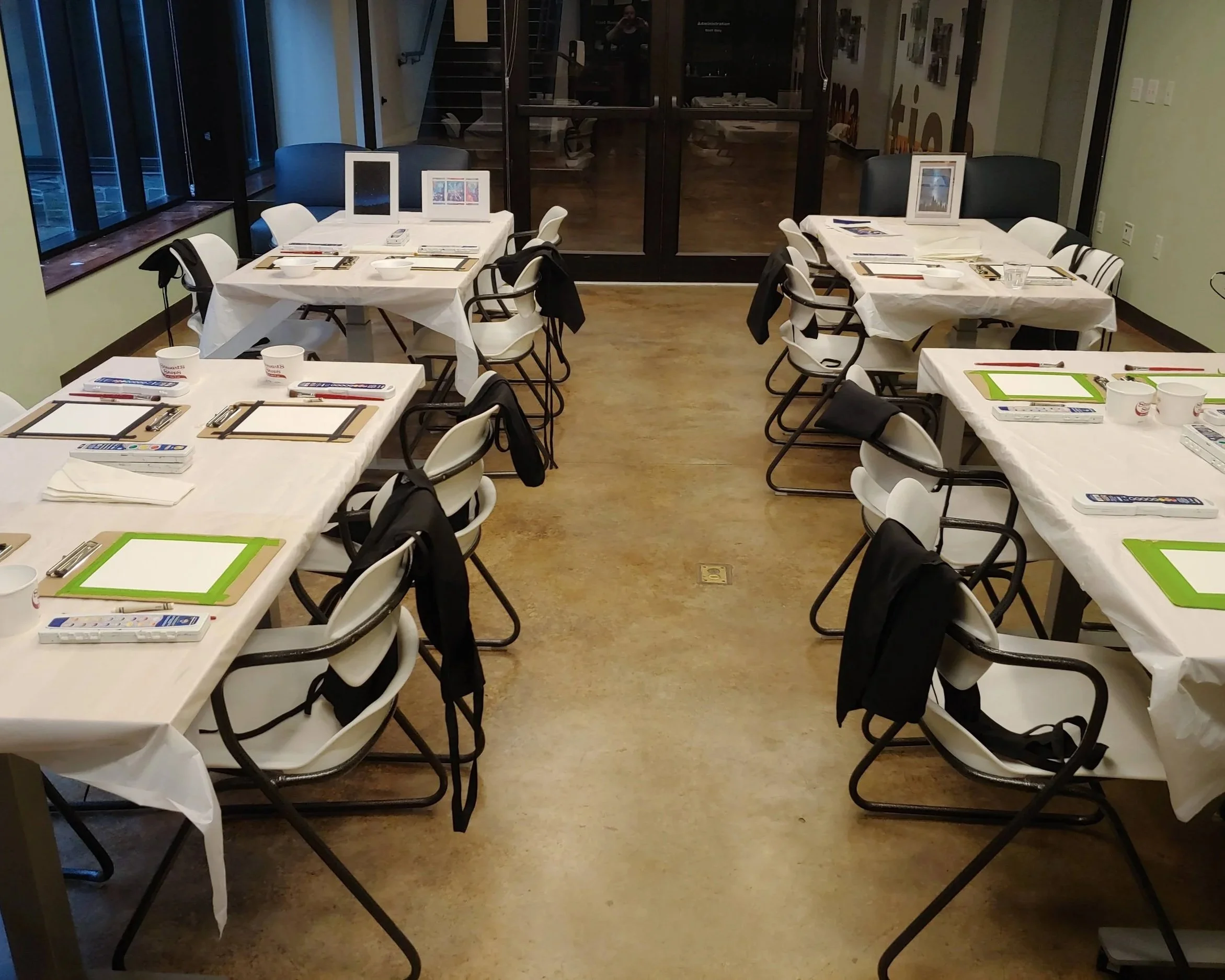 This is an image of the Classroom at the Arkell Museum and Canajoharie Library. There are four tables with chairs and the tables are dressed with white plastic table cloths. There are also watercolor materials (clipboards, paints, water, napkins).