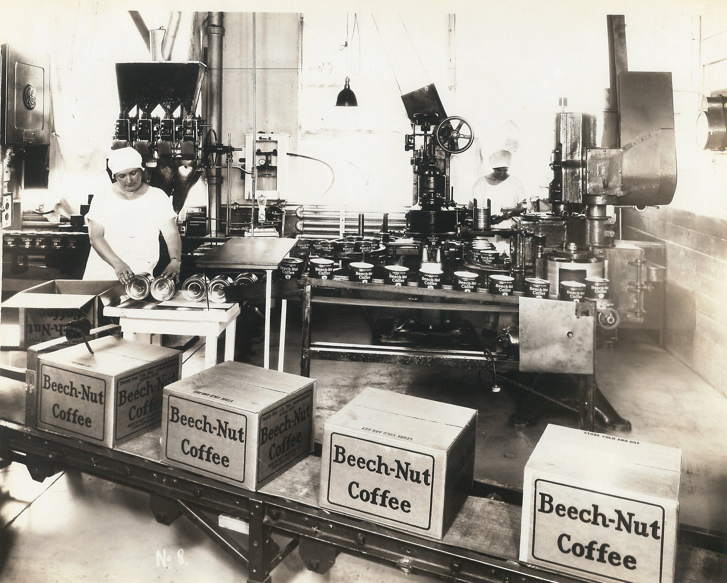 Black and white photo of the interior of a factory where tins of coffee are being placed into boxes. The boxes are marked Beech-Nut Coffee