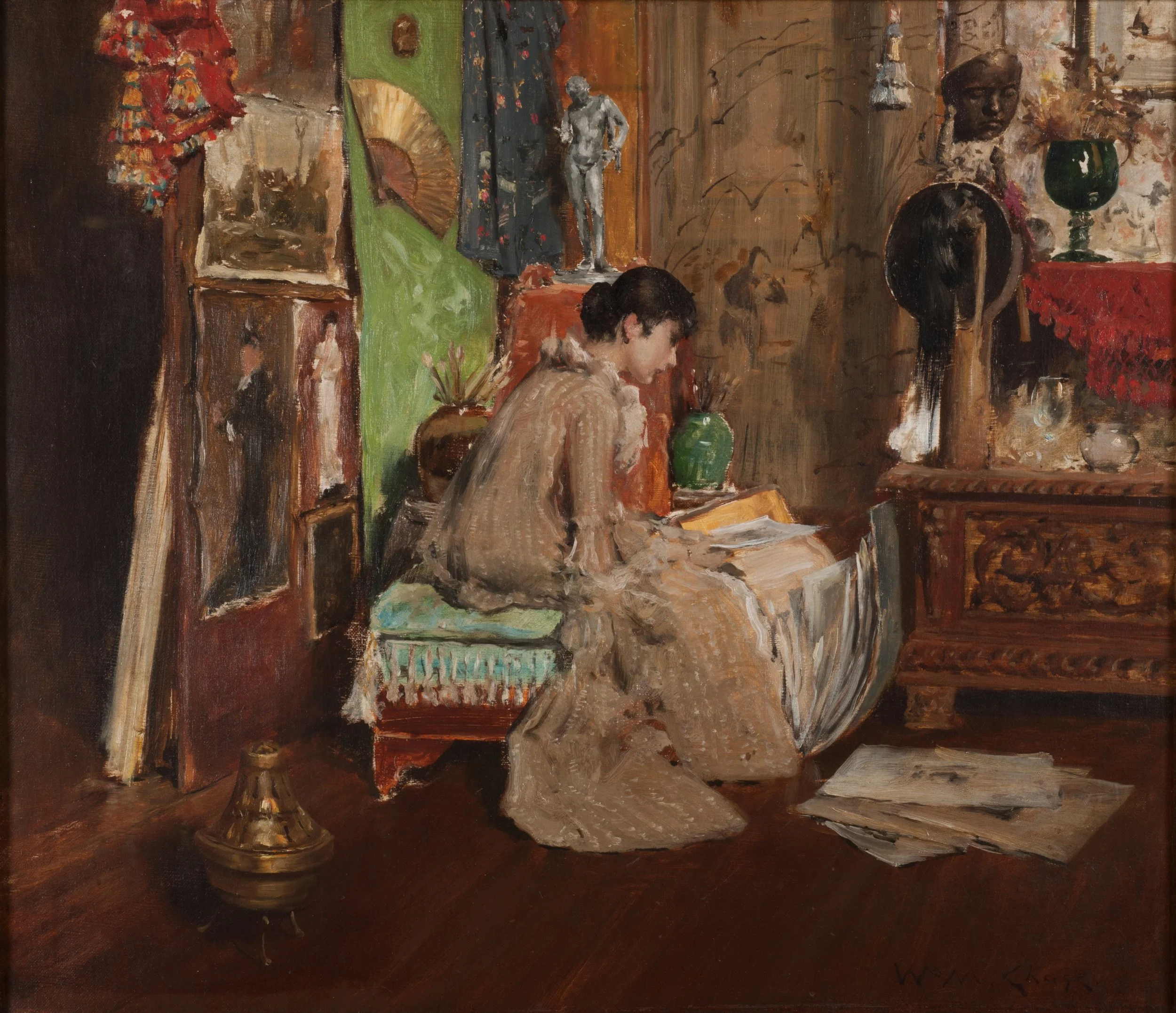 Oil painting of a seated woman who is bent over looking at a stack of paintings. She has selected some any they lay before her feet on the ground. Surrounding her are various paintings and antiques that dress up the artists studio. 