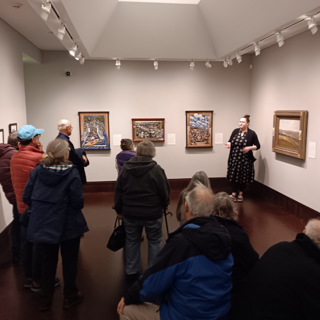 This image features Curator of Education and Public Engagement Mary Alexander providing a tour of a previous exhibition to a group of 10 individuals; Their backs are turned toward the camera, focusing attention on the presentation.