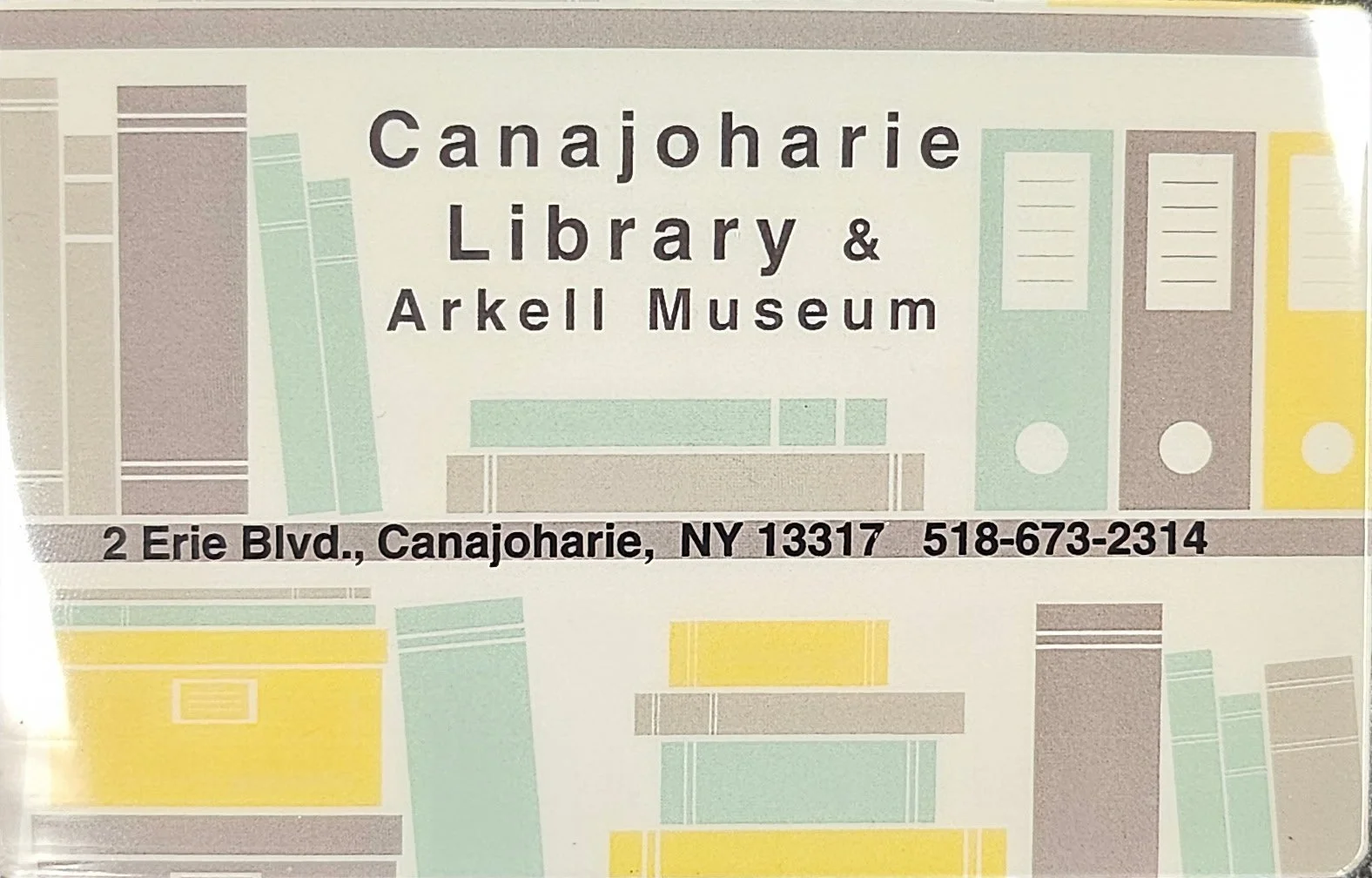This is an image of the Arkell Museum and Canajoharie Library - Library card. It shows our building location and contact details alongside yellow, green, and grey books on a bookshelf.