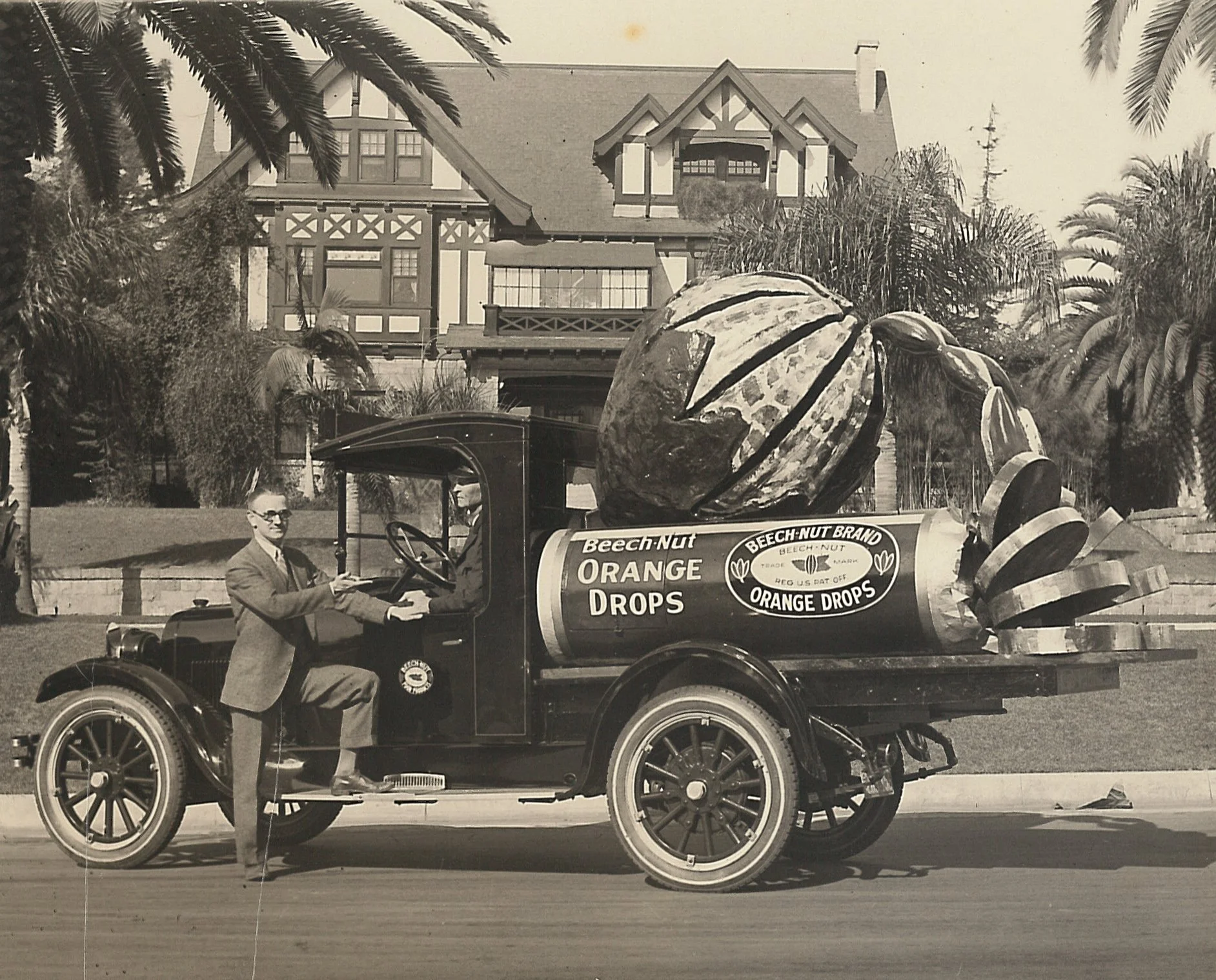 Black and white photo of a truck with a large orange spitting out orange drop candies into a roll wrapper of drops. 