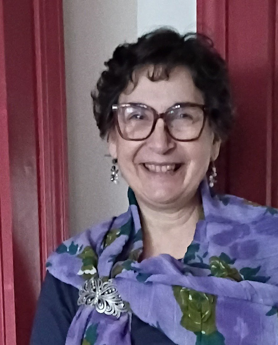 This is a photo of trustee Joanne Resch. She is wearing a purple floral scarf around her neck, dangling earrings, and large glasses.